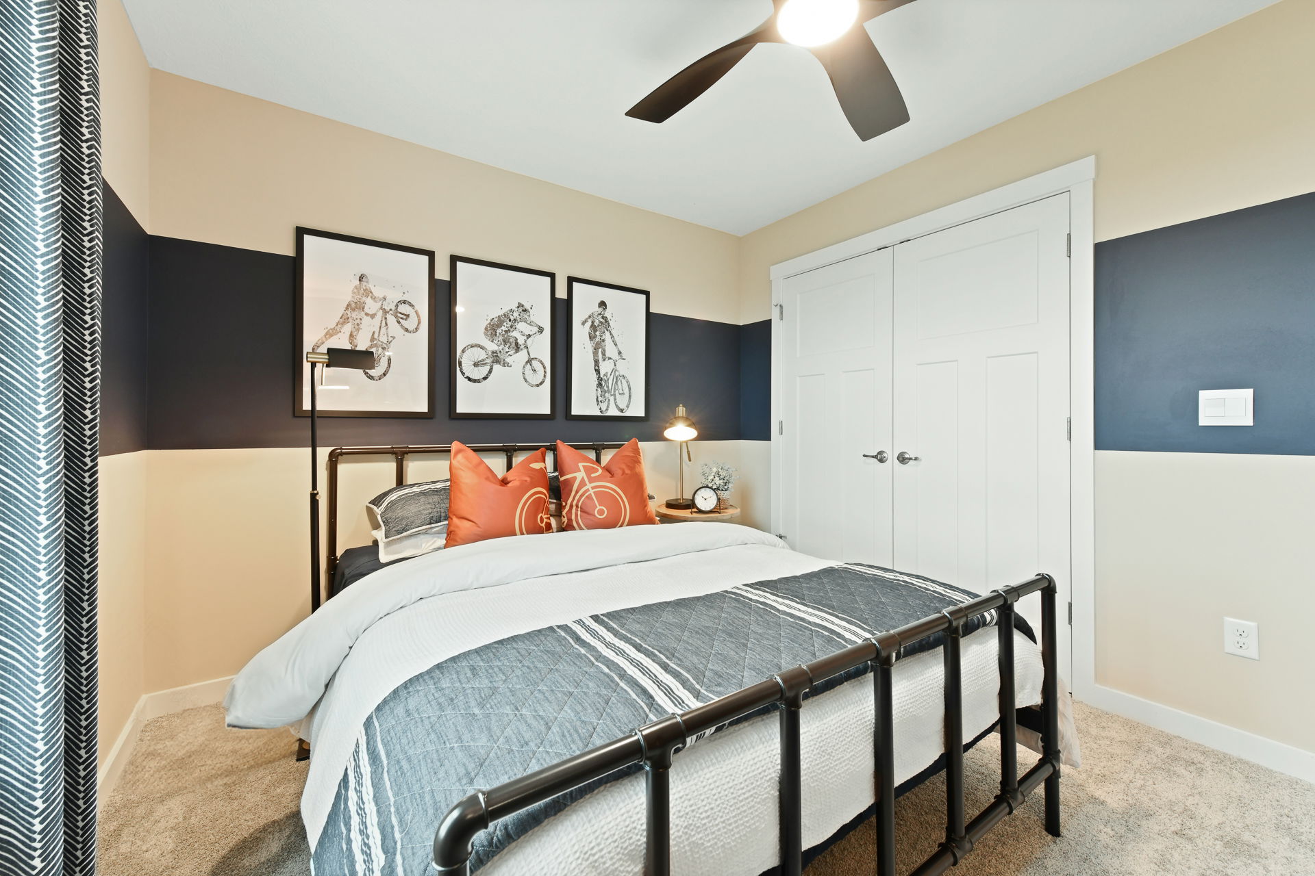 Cozy modern bedroom with beige and navy walls, decorated with motocross artwork and featuring a stylish bed with orange accent pillows and a ceiling fan.