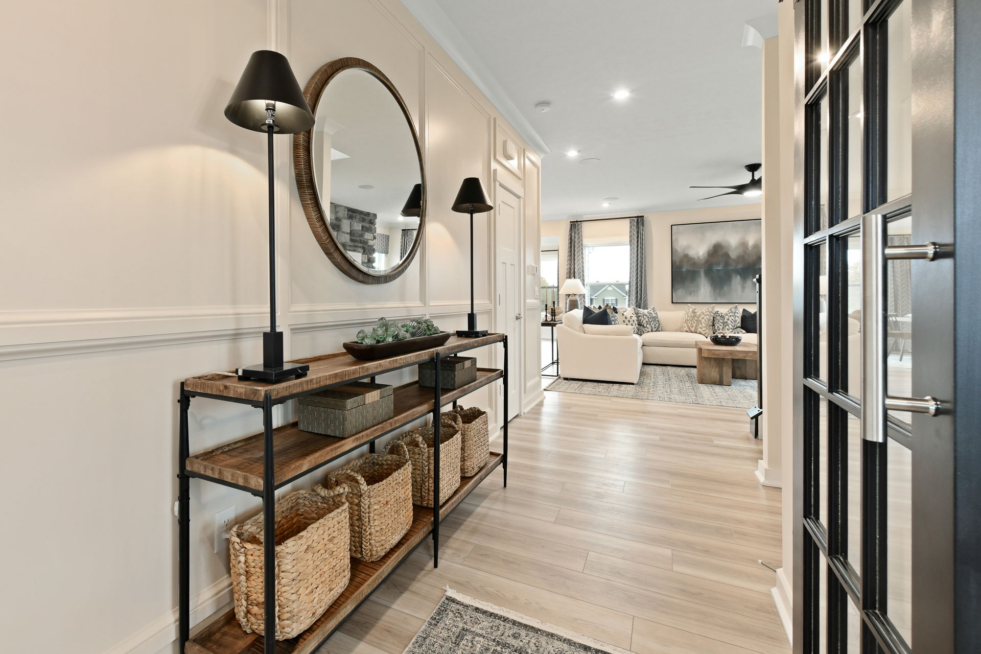Stylish modern entryway with a wooden console table, round mirror, wicker baskets, and lamps leading into a cozy living room with beige seating.