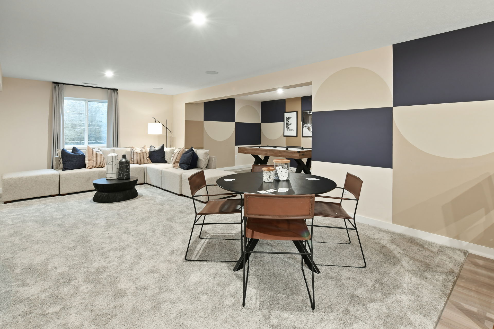 Stylish modern basement game room featuring a cozy beige sectional, round table with leather chairs, and geometric wall art.