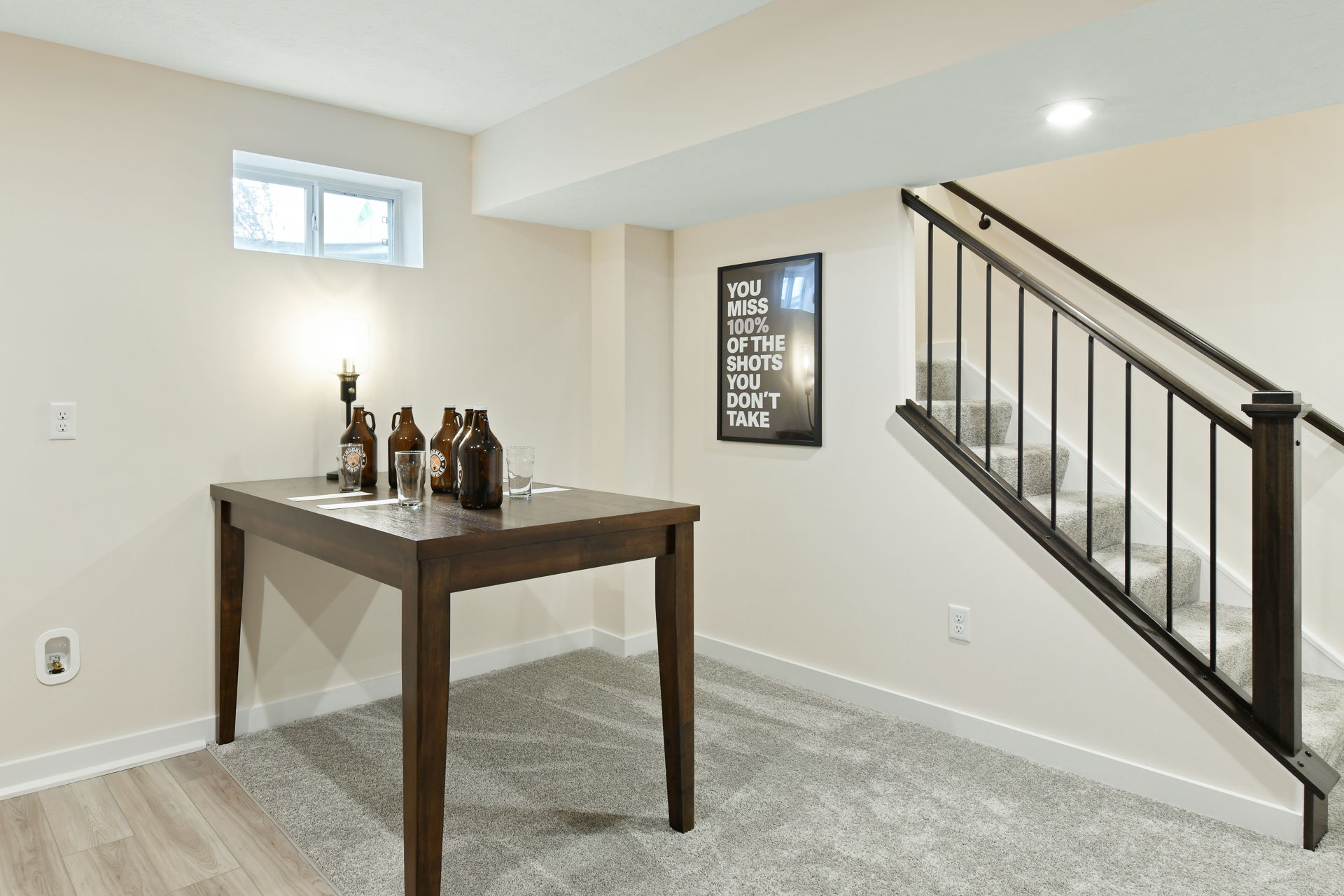 Modern basement room with a dark wooden table, decorative bottles, a framed quote art, and carpeted stairs.