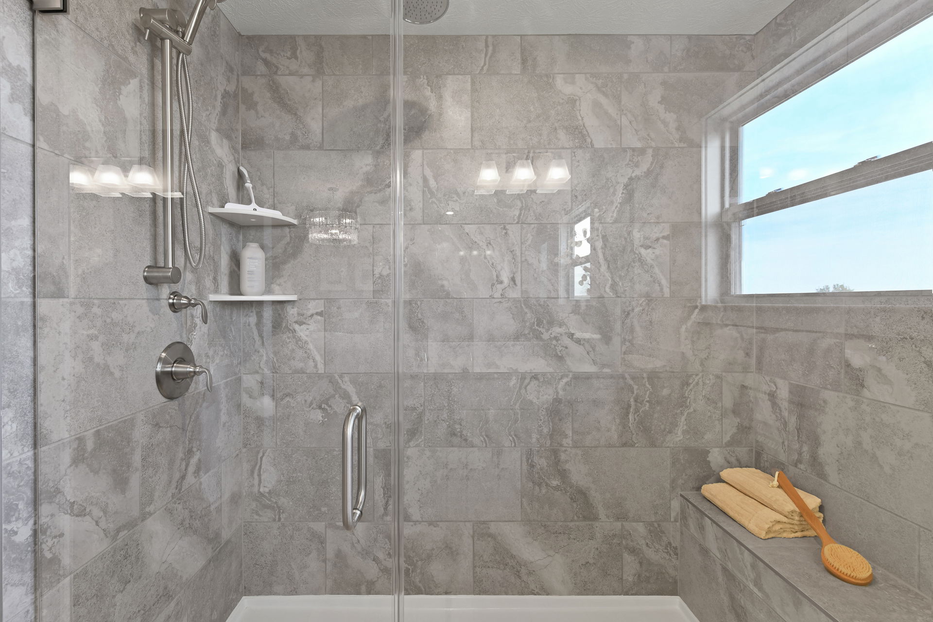 Modern bathroom shower with sleek gray tiles, a glass door, built-in shelves, and a window offering natural light.