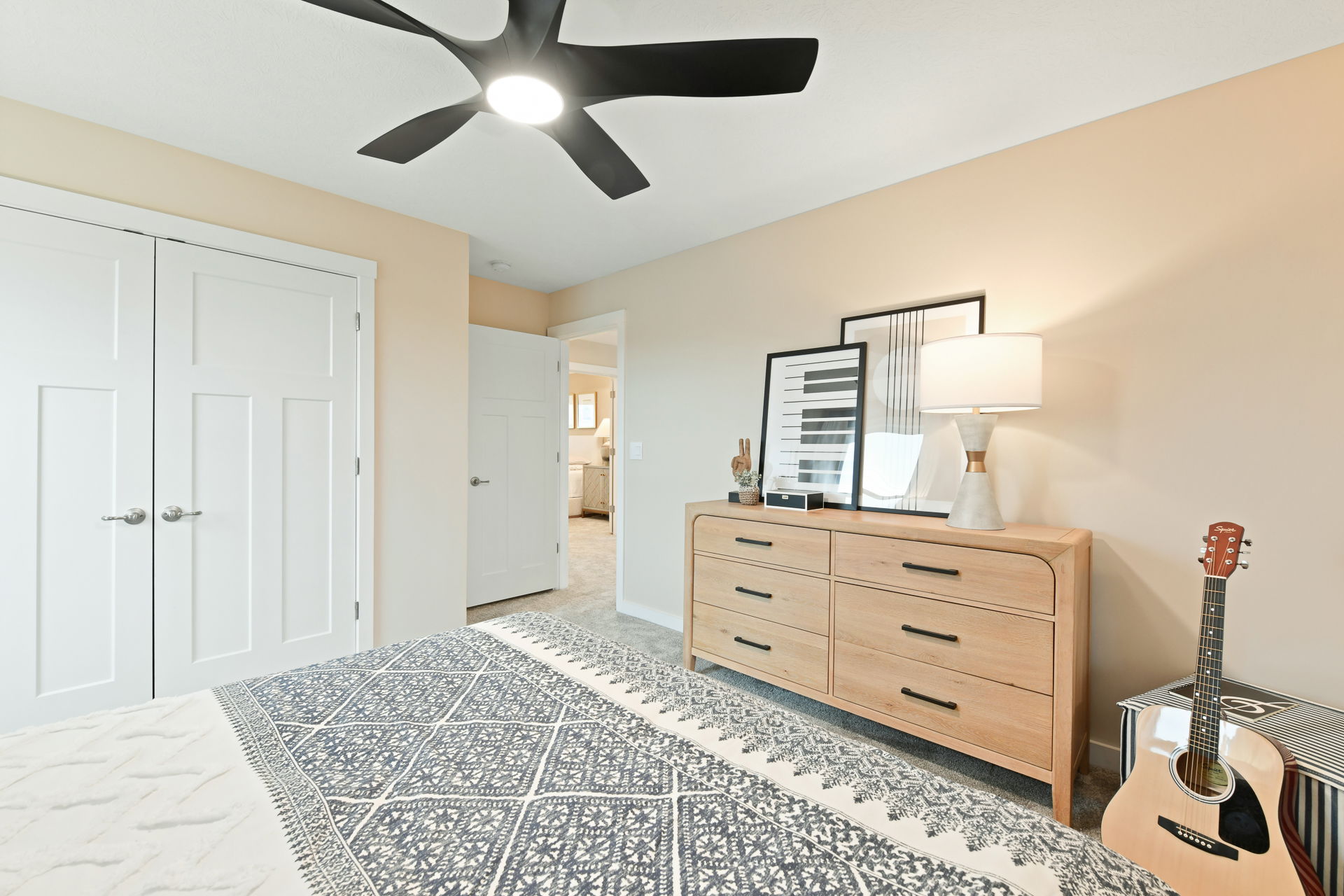 Modern bedroom featuring a stylish wooden dresser, decorative lamp, patterned bedding, acoustic guitar, and contemporary ceiling fan.