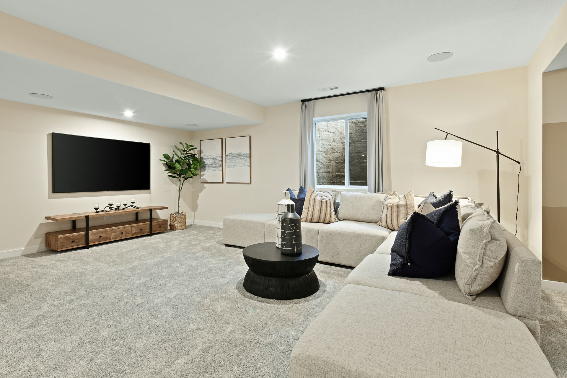 Modern basement living room with a large sectional sofa, flat-screen TV, stylish decor, and natural light from the window.