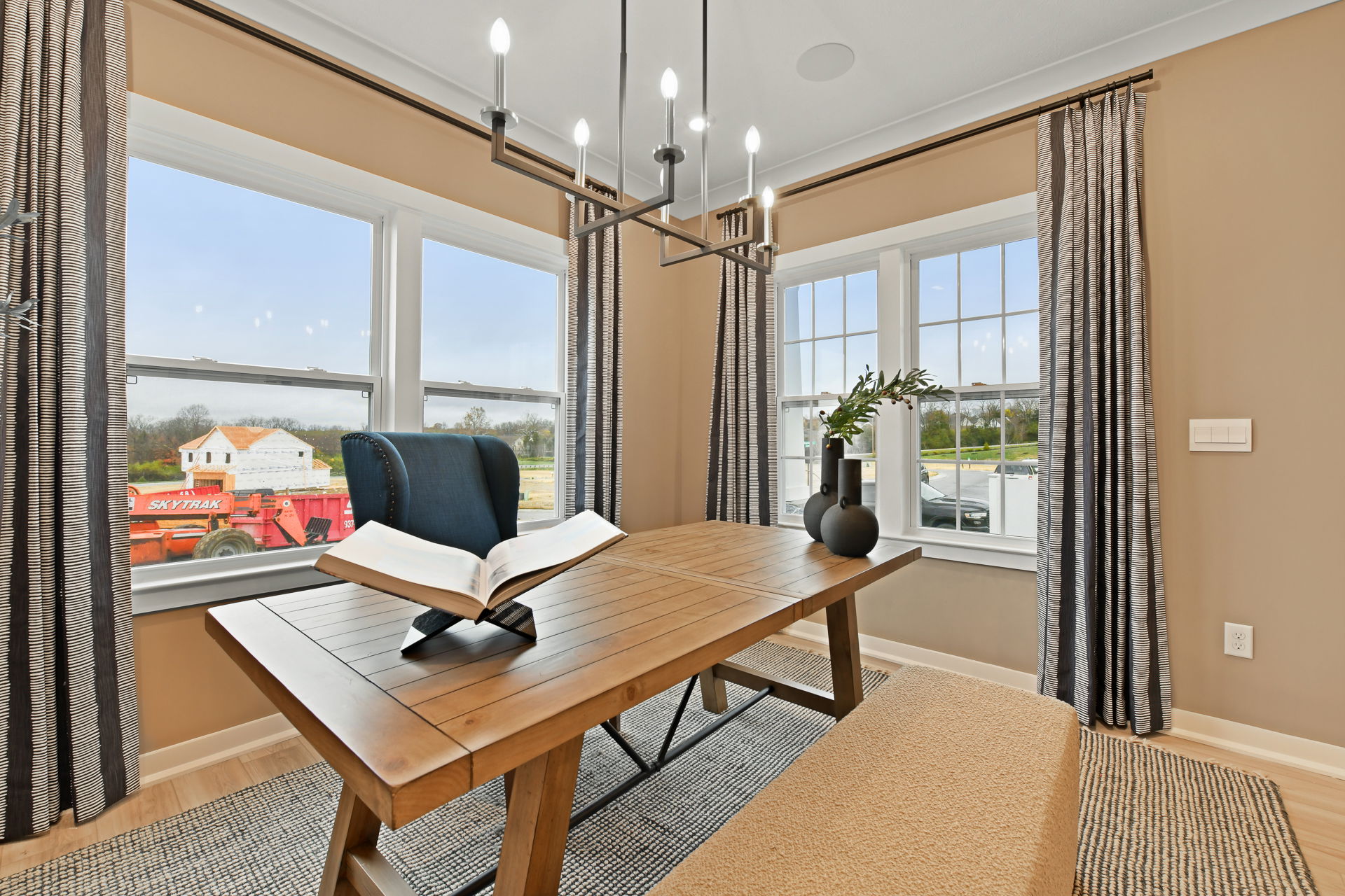 A modern dining room with a wooden table, a blue upholstered chair, a book on a stand, and large windows showcasing an outdoor construction scene.