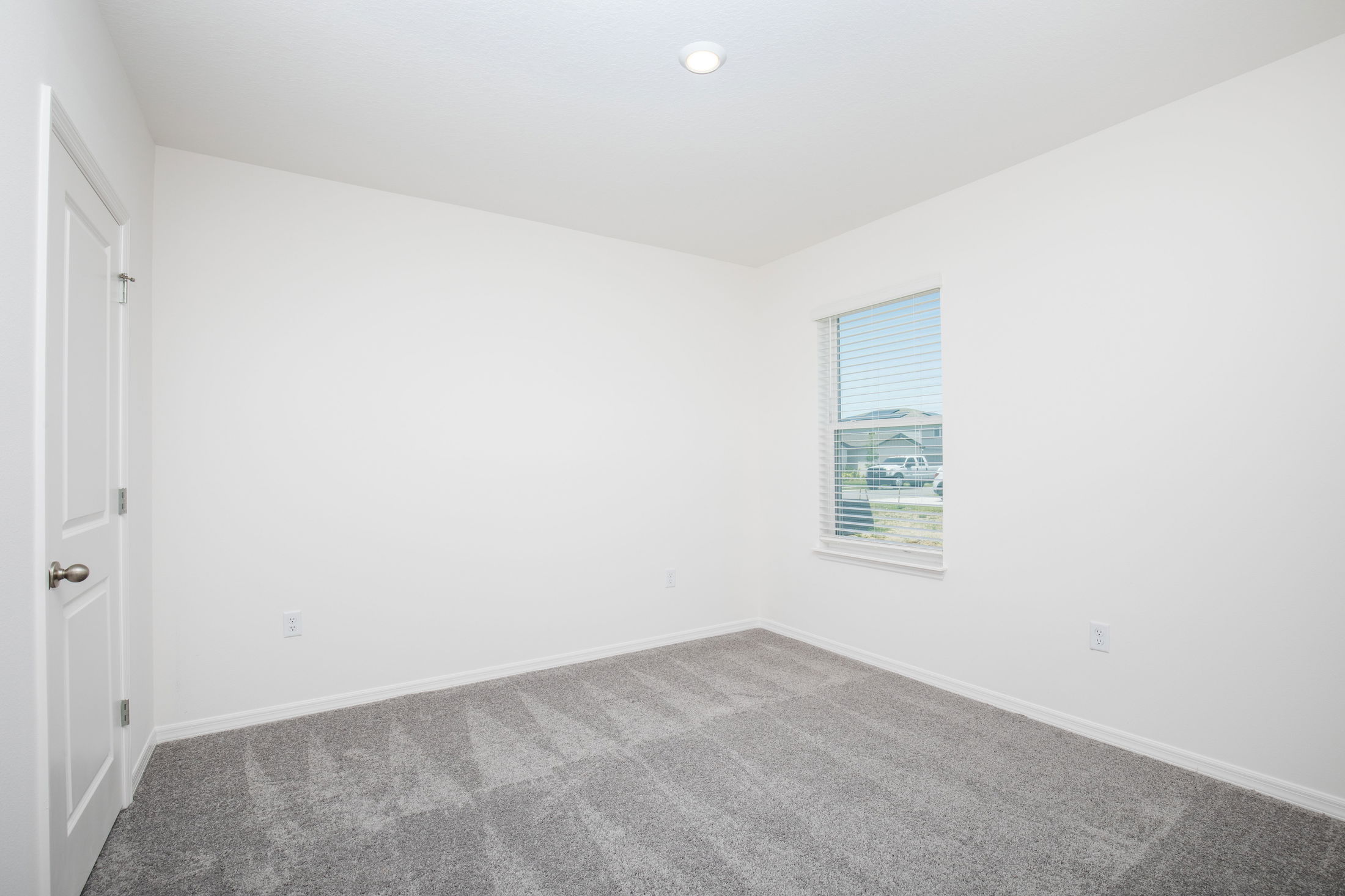 Bright, empty room with white walls, a window with blinds, and clean gray carpeting.