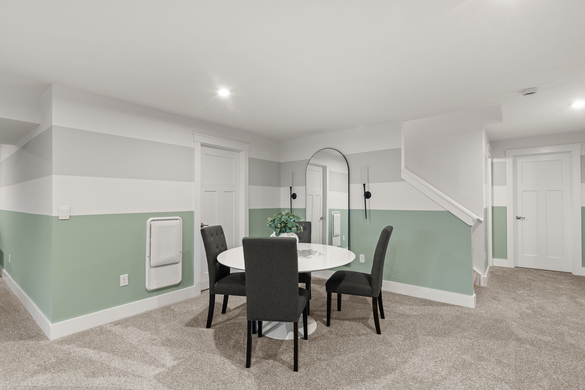 Modern basement dining area with a round white table, four dark upholstered chairs, and stylish two-tone walls.