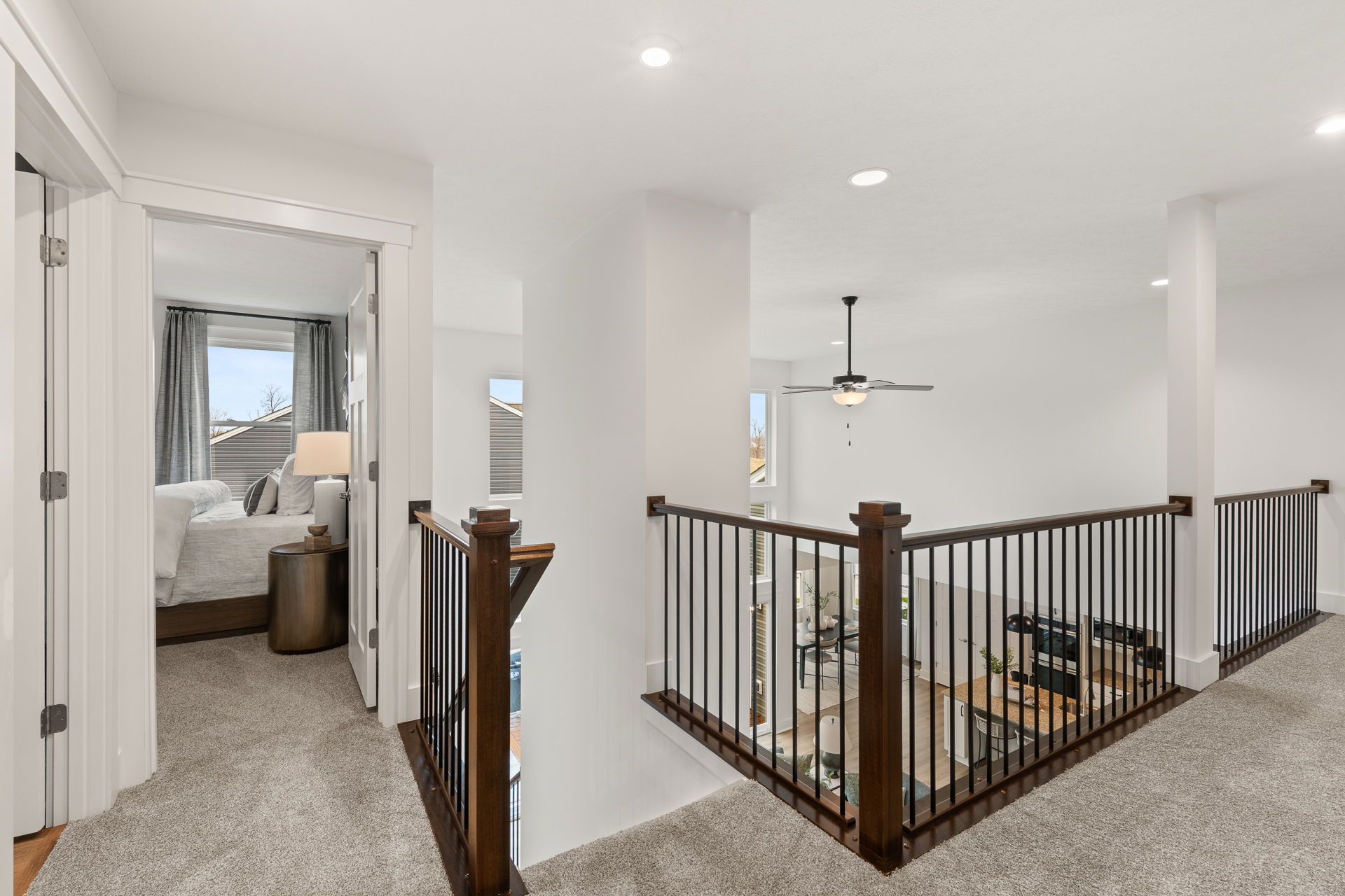 Modern home interior with open upstairs hallway, metal and wood railing, leading to a furnished bedroom and overlooking the living area with large windows and a ceiling fan.