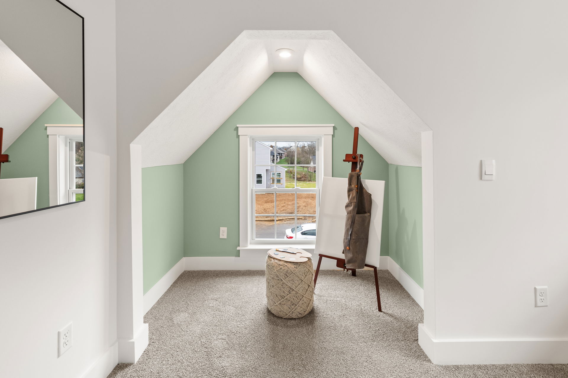 Cozy attic art studio with a window, featuring a canvas on an easel and a woven ottoman against green and white walls.
