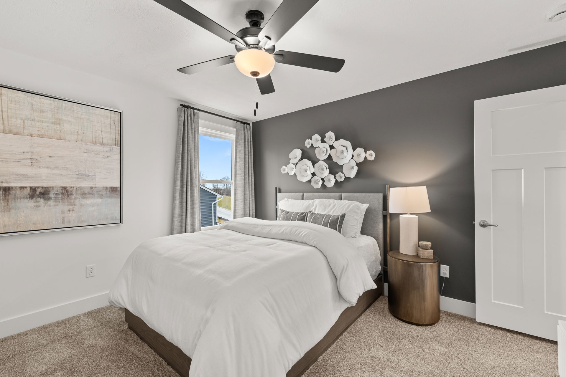 Modern bedroom with gray accent wall, elegant white bedding, and decorative wall flowers, featuring natural light from a large window.