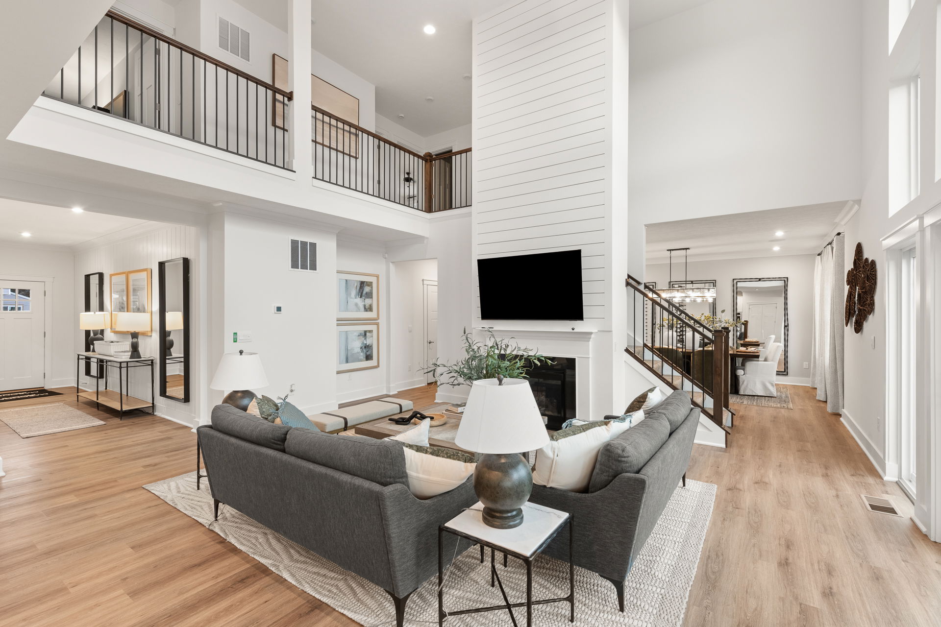 Spacious modern living room with high ceilings, featuring a gray sectional sofa, wall-mounted TV above a fireplace, and wooden flooring.