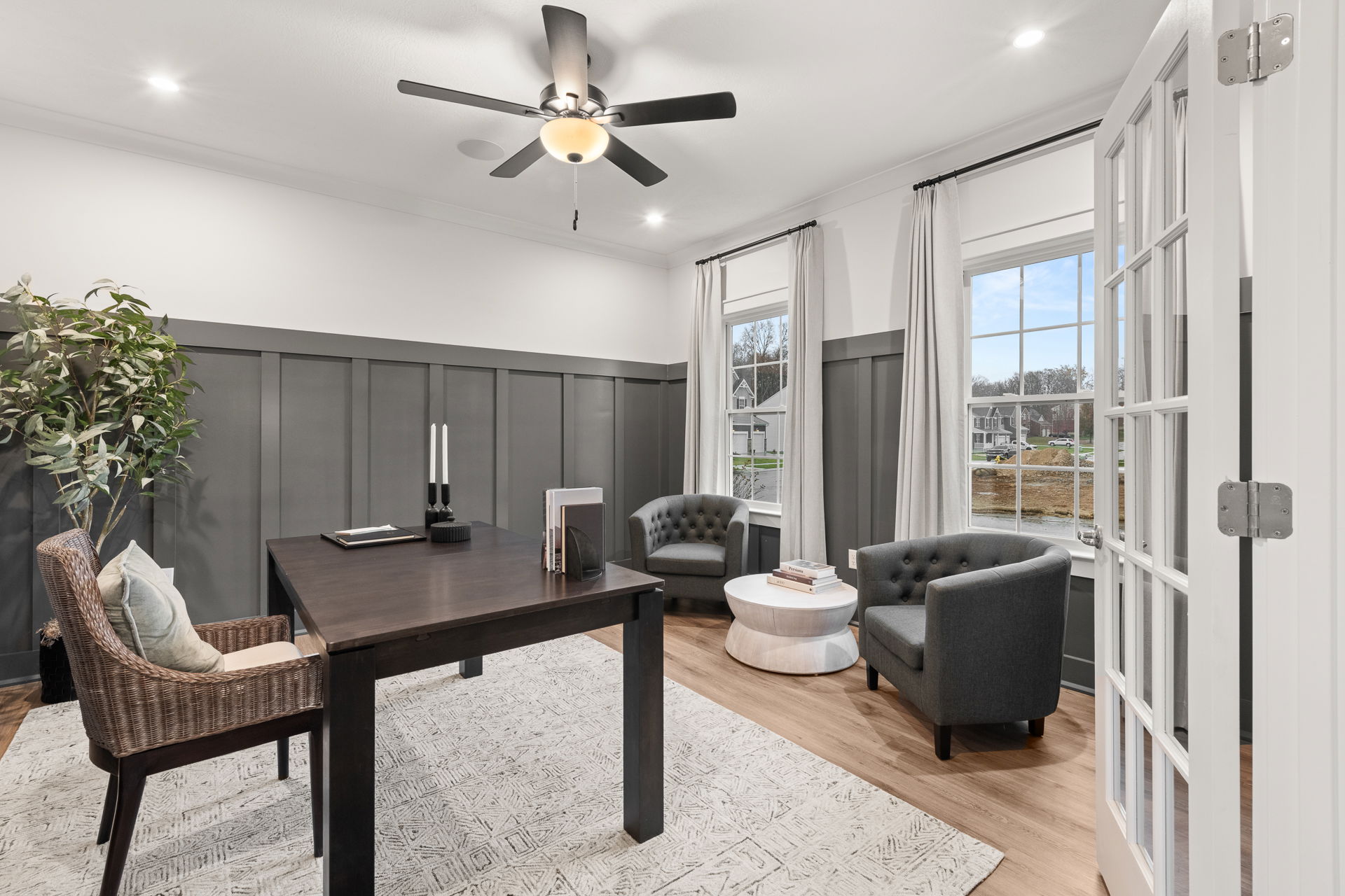 Modern home office with a dark wood desk, wicker chair, two gray armchairs, and large windows for natural light.