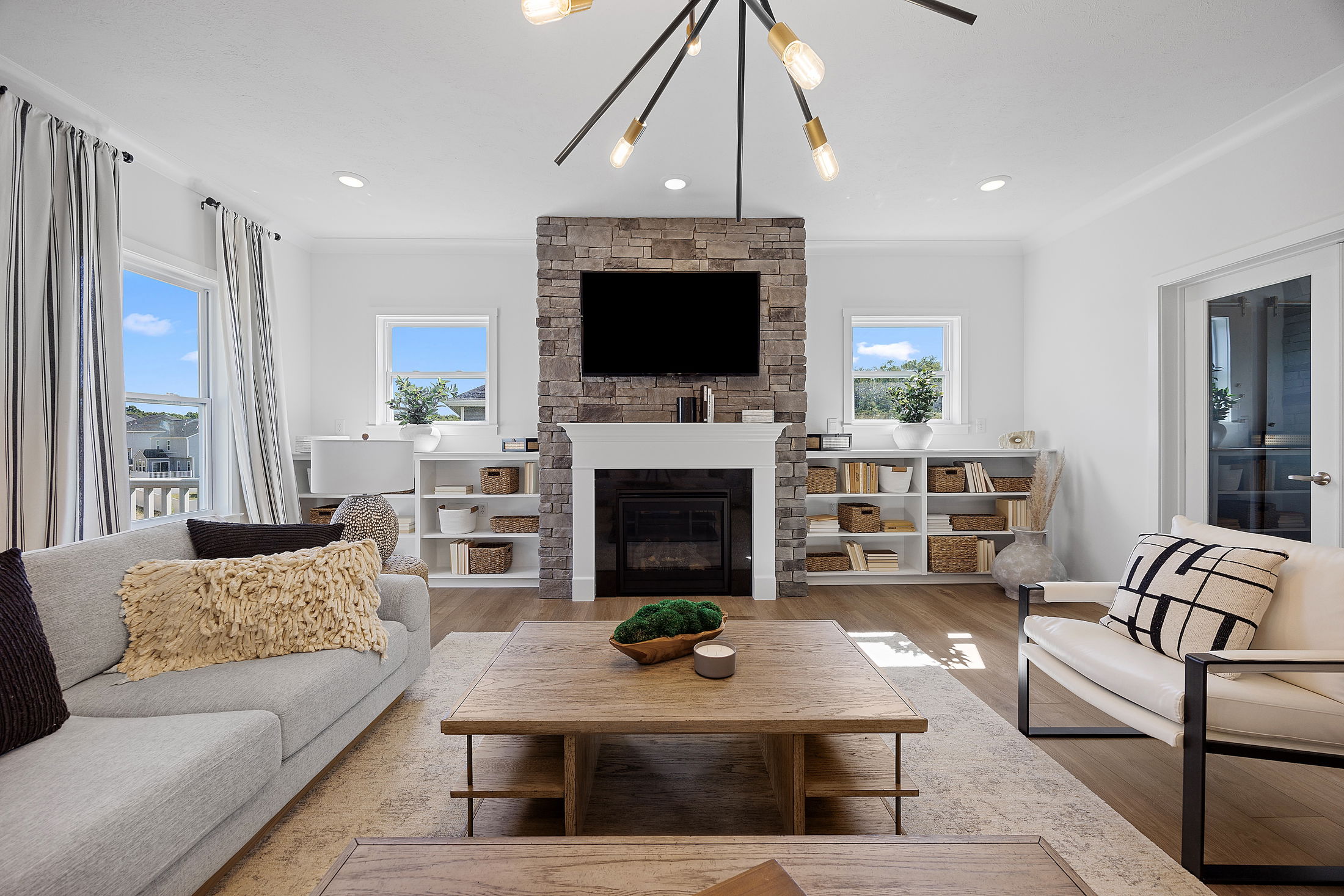 Modern living room with a cozy gray sofa, stone fireplace, and decorative bookshelves, featuring large windows and contemporary lighting.