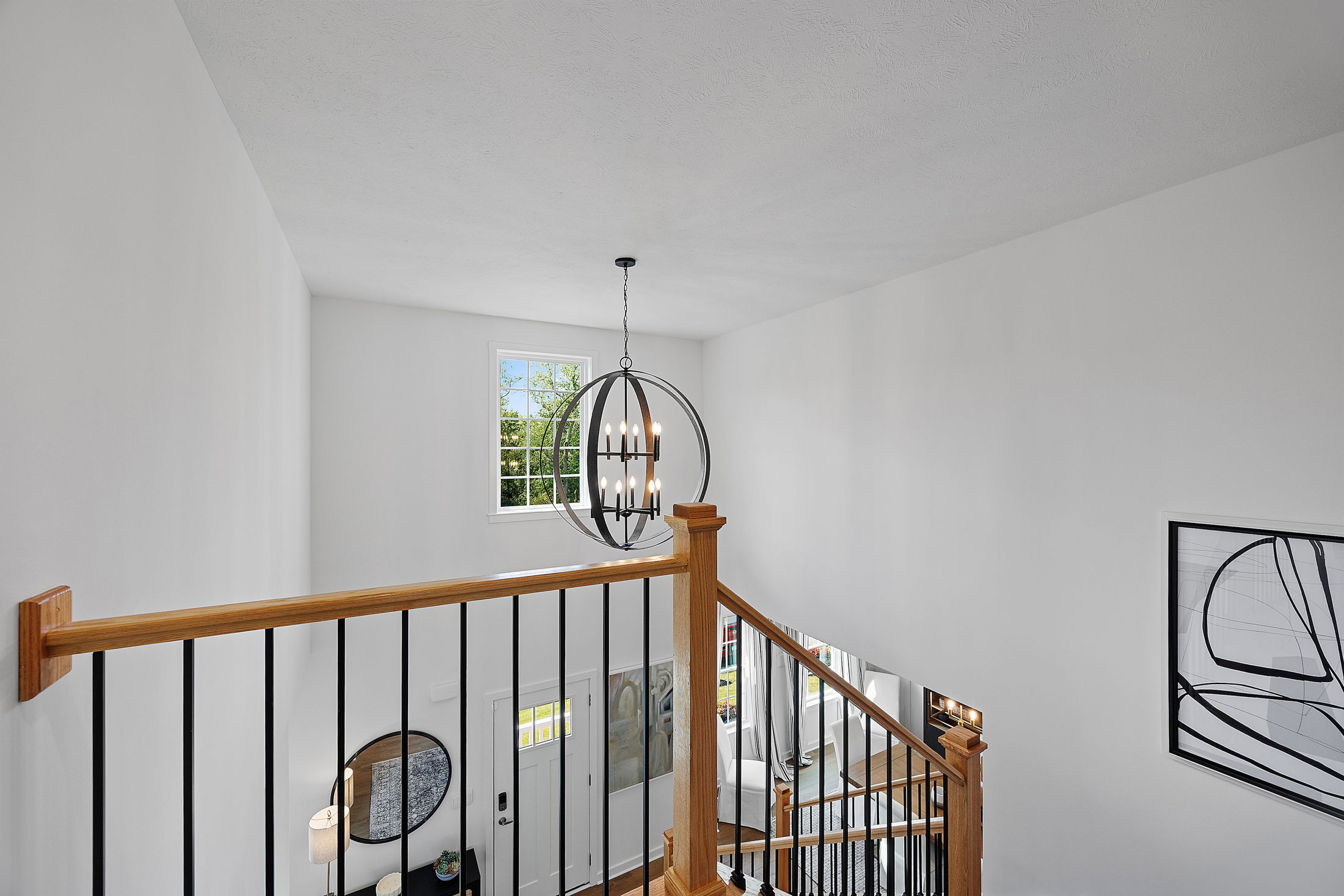 Contemporary staircase with wooden railings and black spindles, illuminated by a spherical chandelier near a large window.