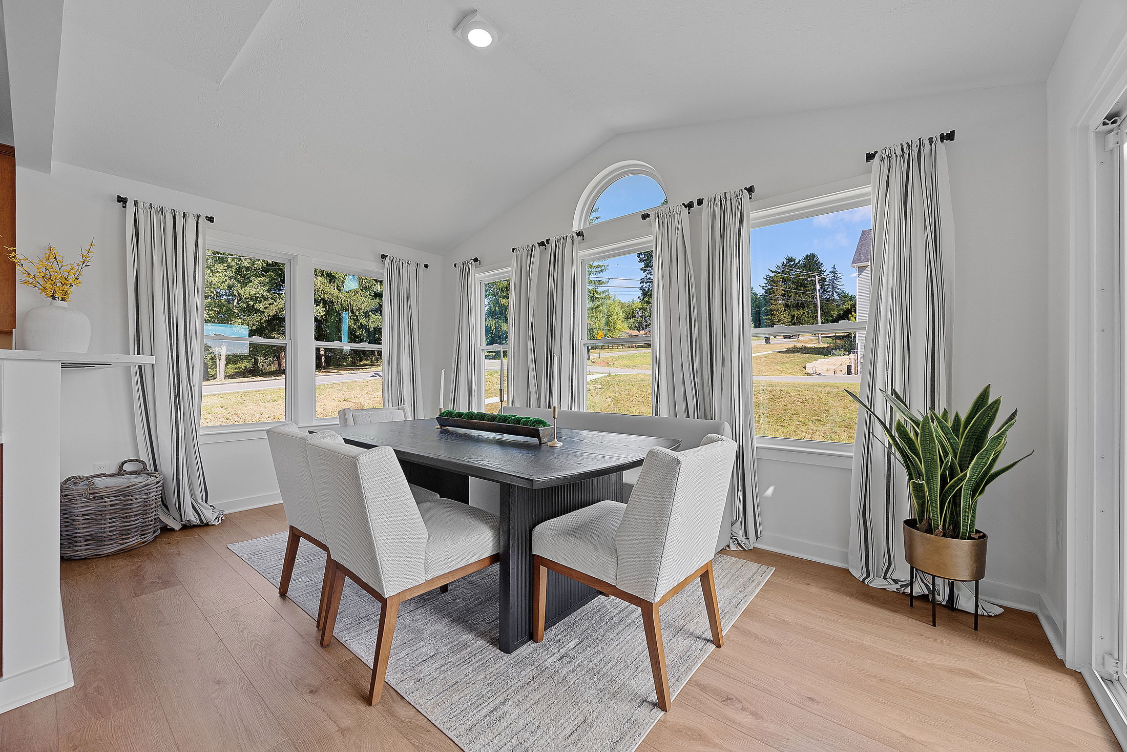 Bright, modern dining room with a dark wood table, white chairs, large windows, and green indoor plant.