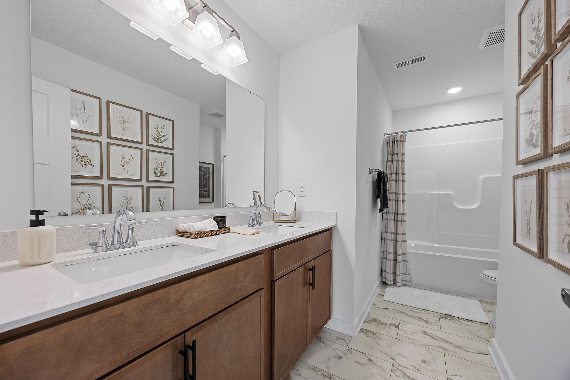 Modern bathroom interior with a double vanity, framed botanical prints, and a shower-tub combo.