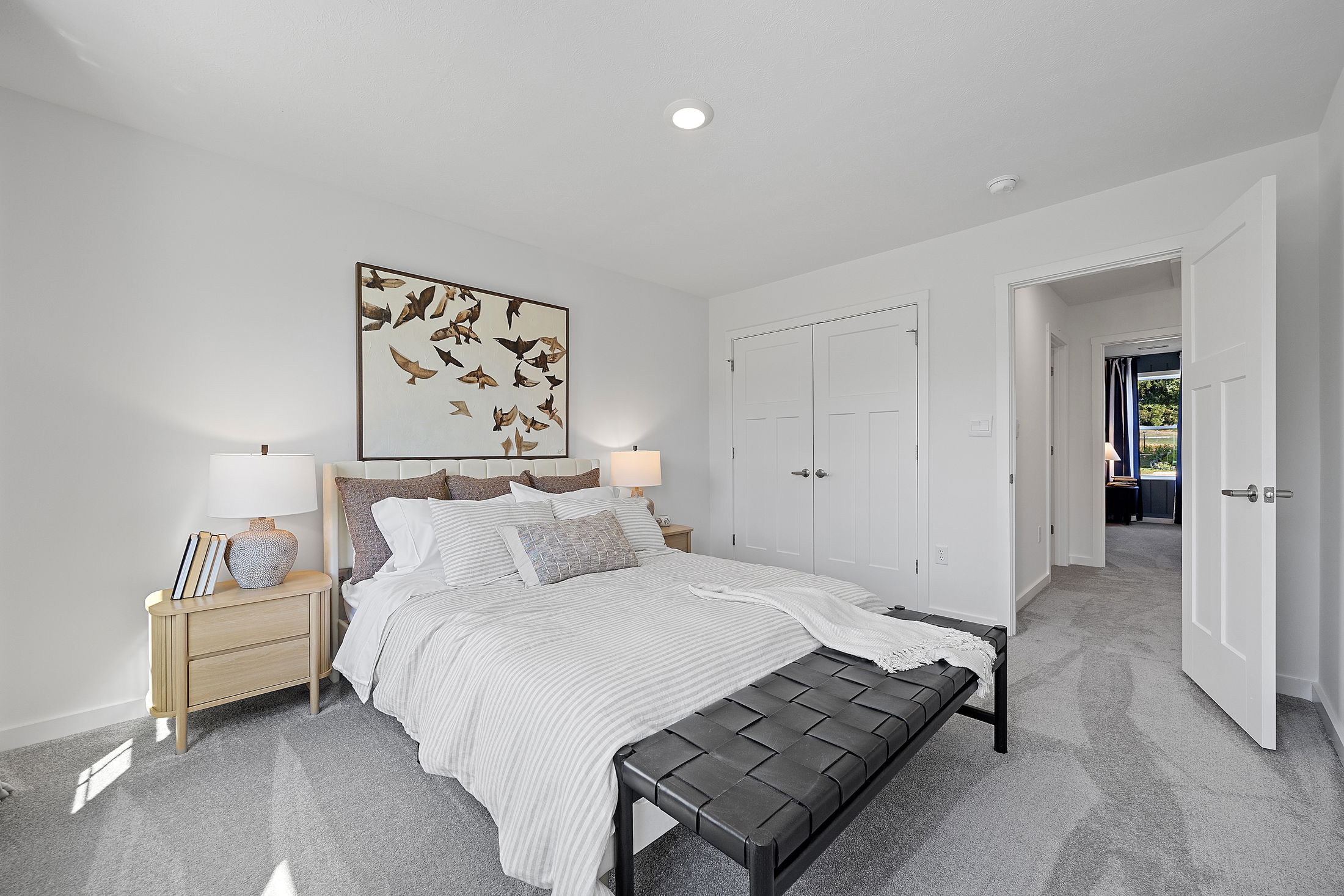 Modern bedroom with a cozy white bed, wooden nightstands, bird-themed artwork, and open doors leading to a bright hallway.