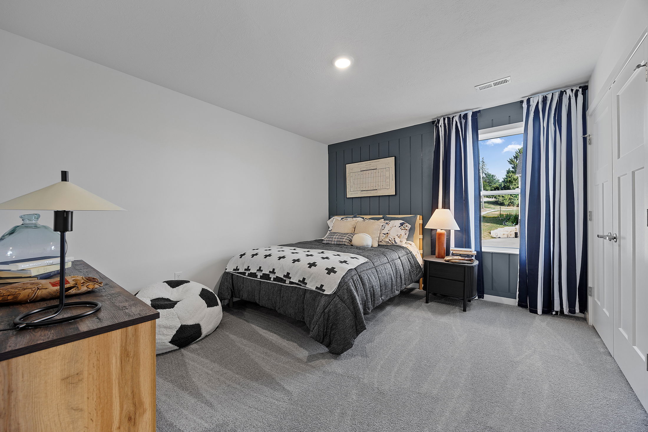 Modern bedroom with gray and black bedding, striped curtains, a bedside lamp, and a soccer ball bean bag chair on a carpeted floor.