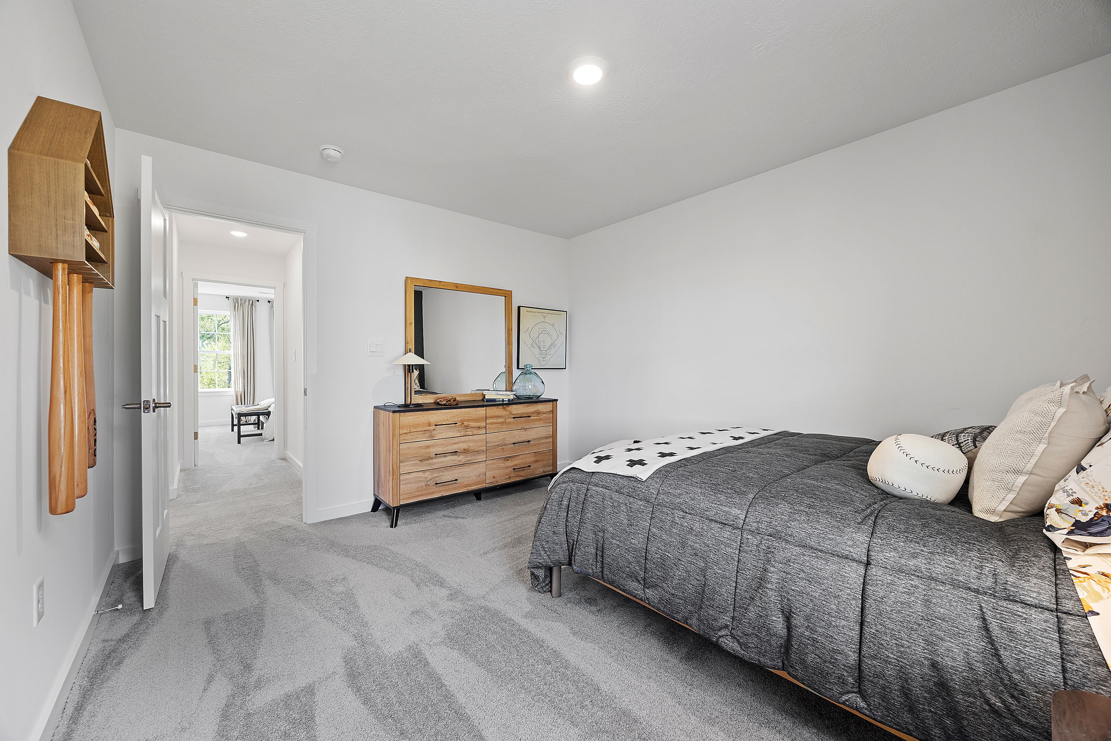 Modern bedroom with gray carpet, wooden dresser, and cozy bedding, leading to a sunlit room with an open door.