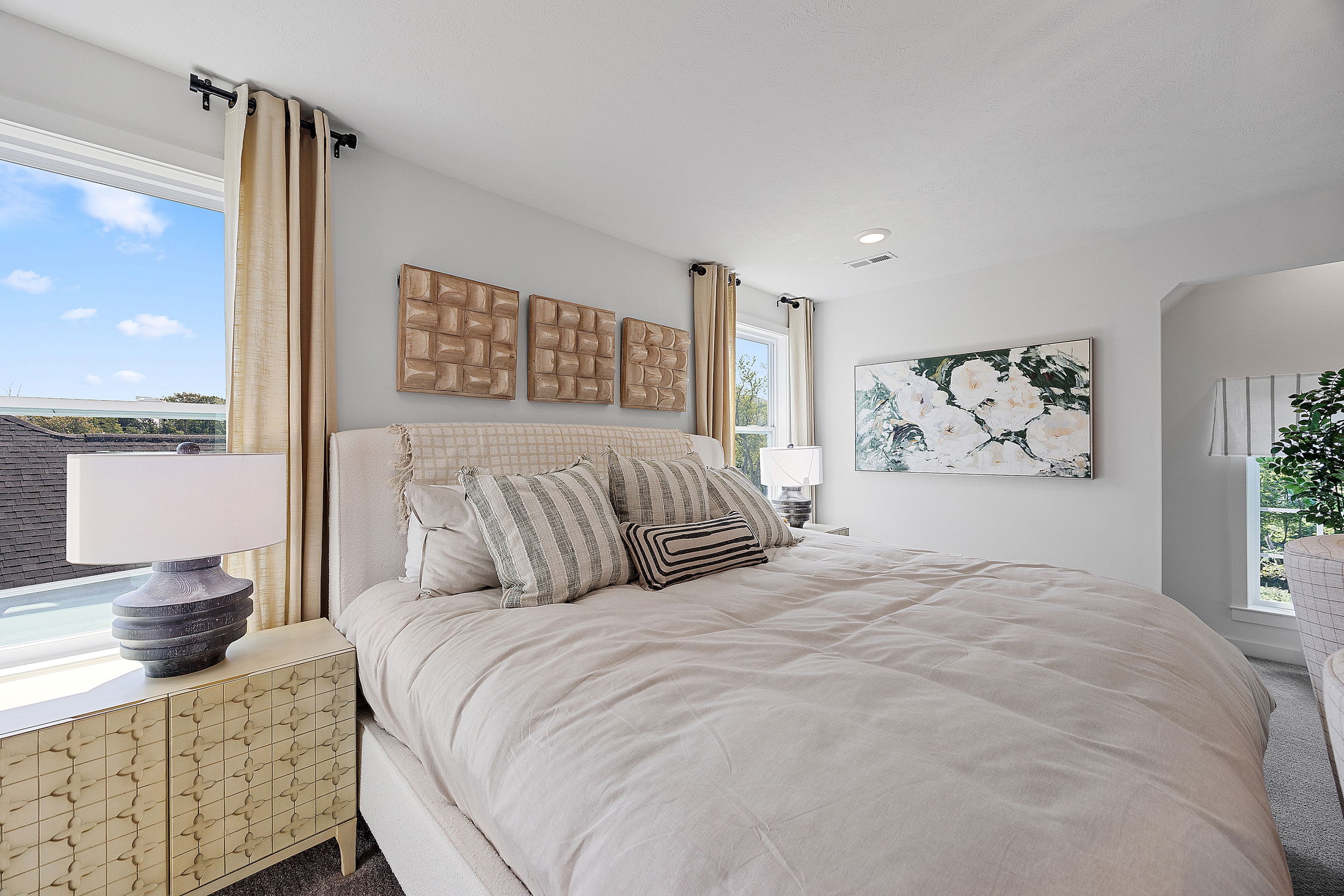 Modern bedroom featuring elegant beige decor, plush bedding with striped pillows, and artistic wall art near large windows.