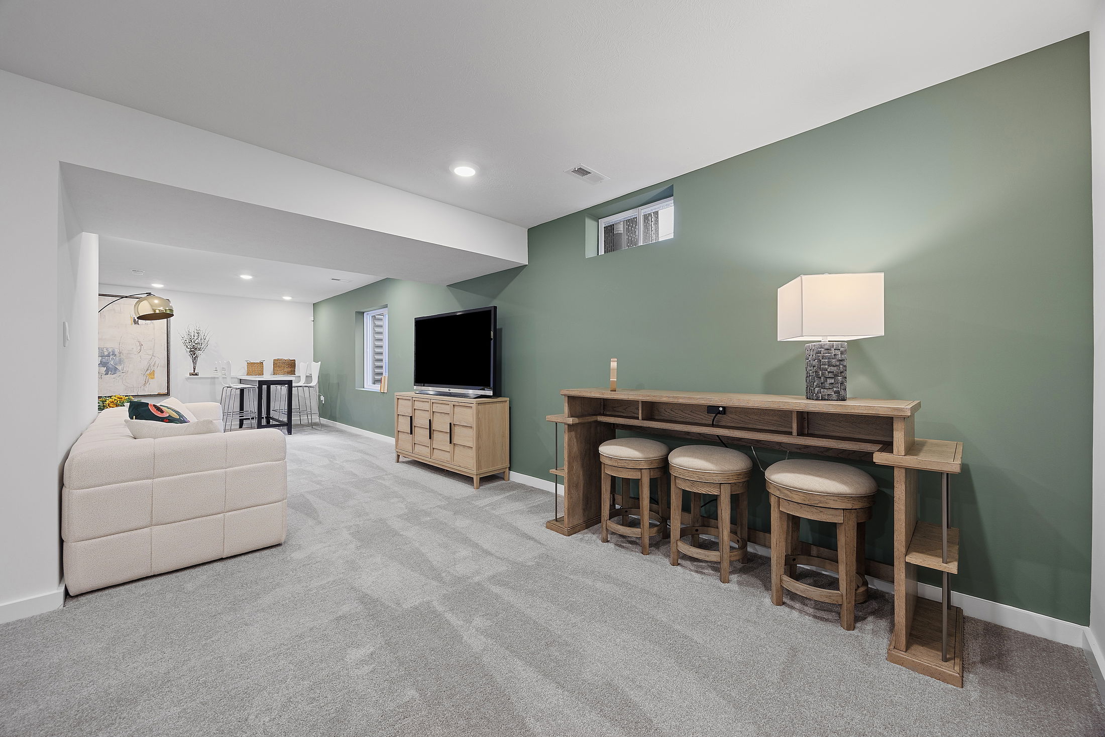 Modern basement entertainment room with green accent wall, comfortable beige seating, wooden furniture, a TV, and a stylish bar area.