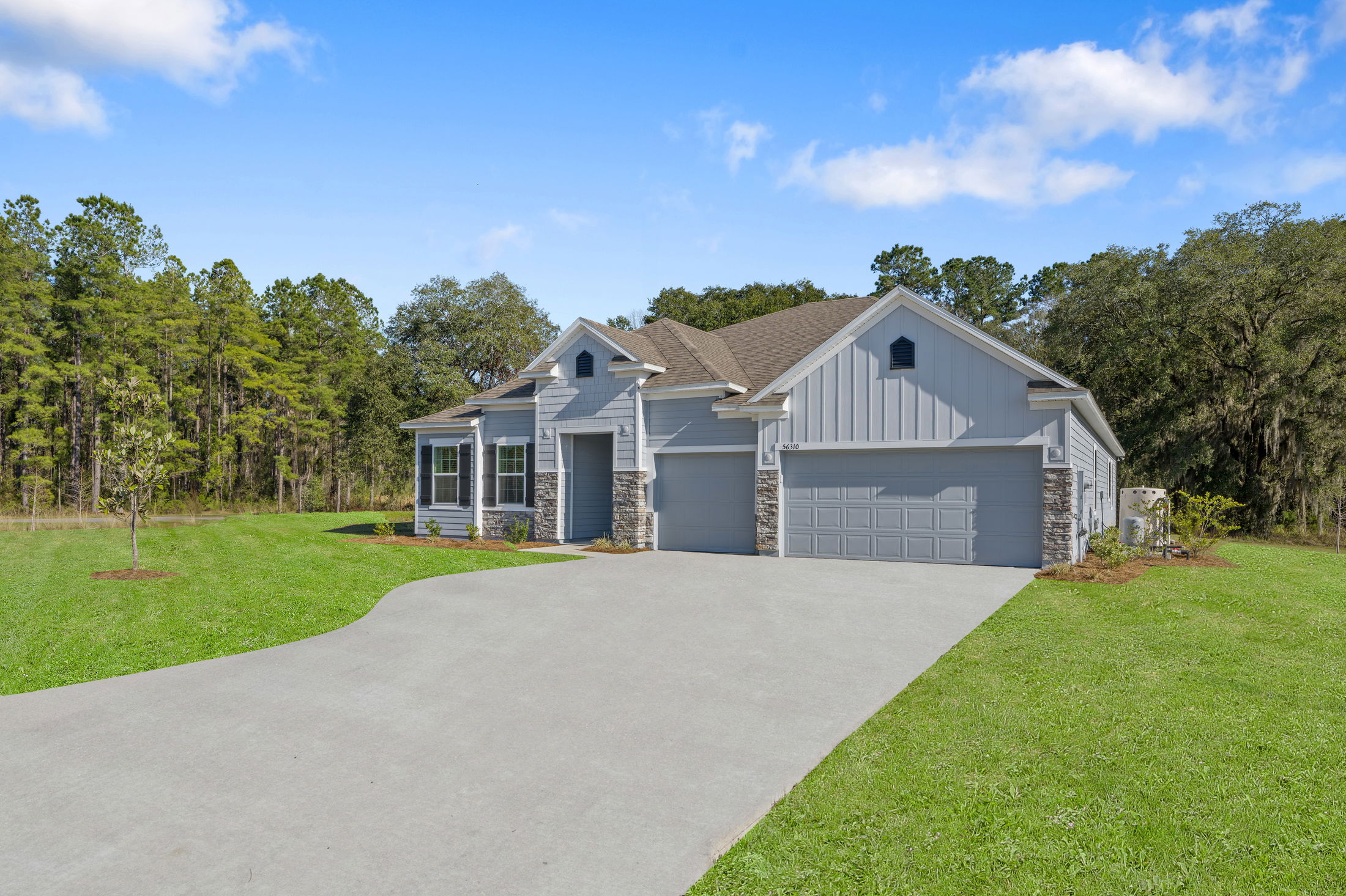 A modern, single-story gray house with stone facade detailing, surrounded by lush greenery and a spacious driveway, set against a backdrop of tall trees and a clear blue sky.