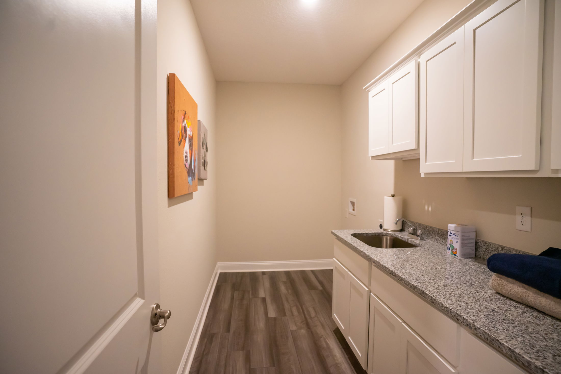 Laundry room with granite countertop, white cabinets, and wall art, featuring hardwood floors and a stainless steel sink.