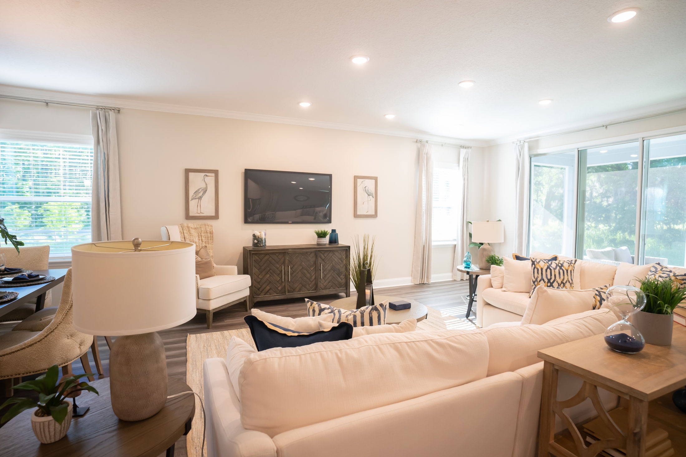 Modern living room with white couches, large windows, and wall-mounted TV, decorated with blue accents and nature-themed artwork.