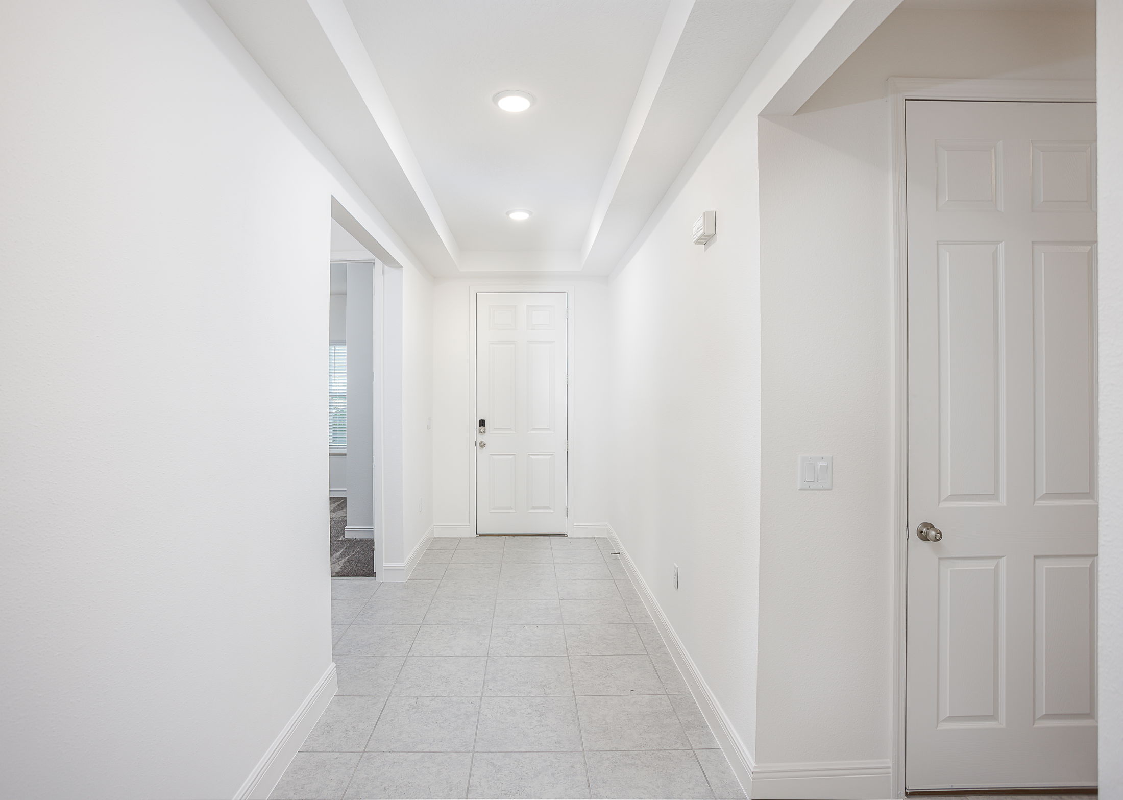 Minimalist hallway with white walls, recessed lighting, and tiled floor leading to a closed white door.