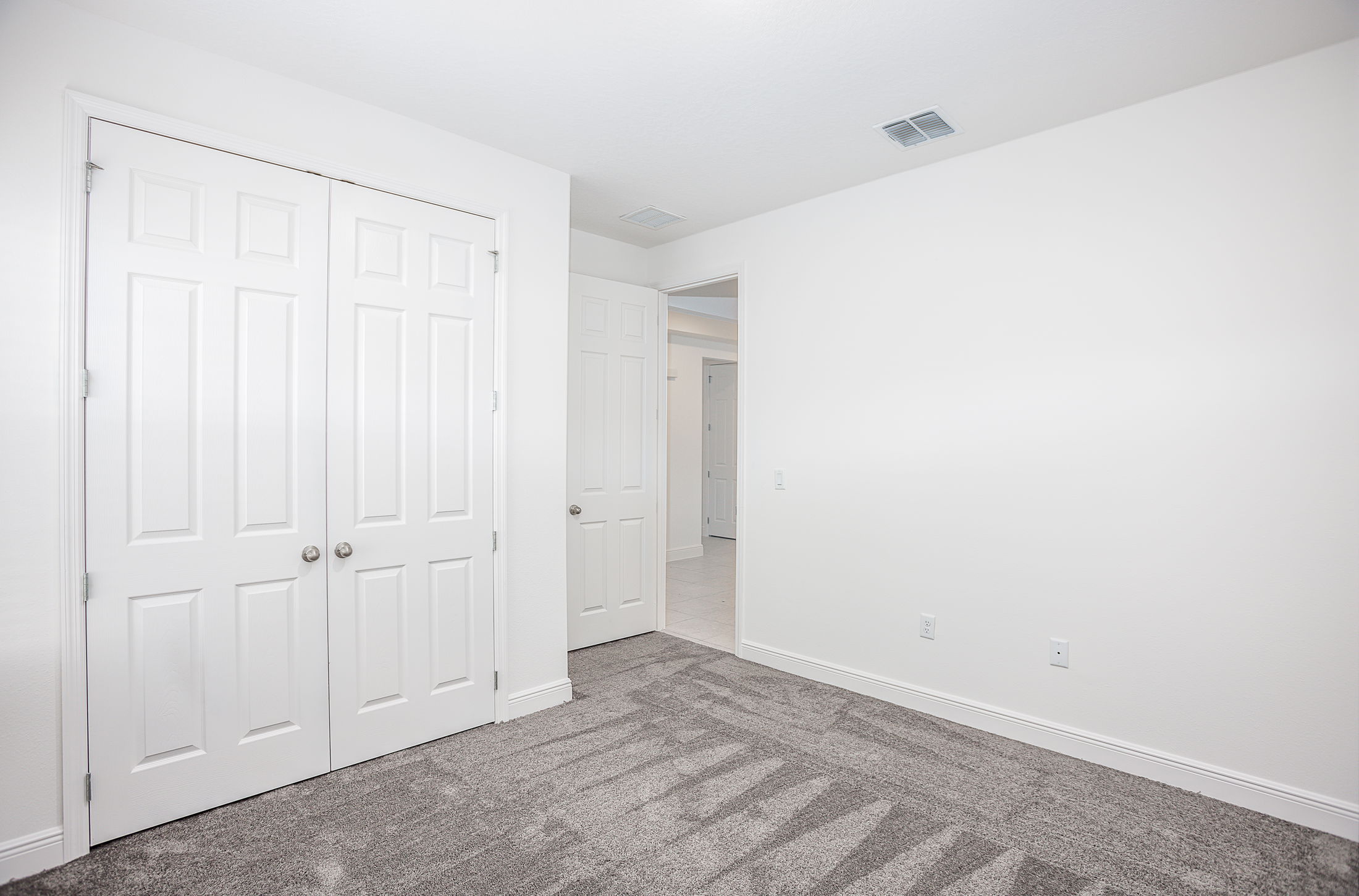 Empty white room with gray carpet, double closet doors, and an open door leading to a hallway.