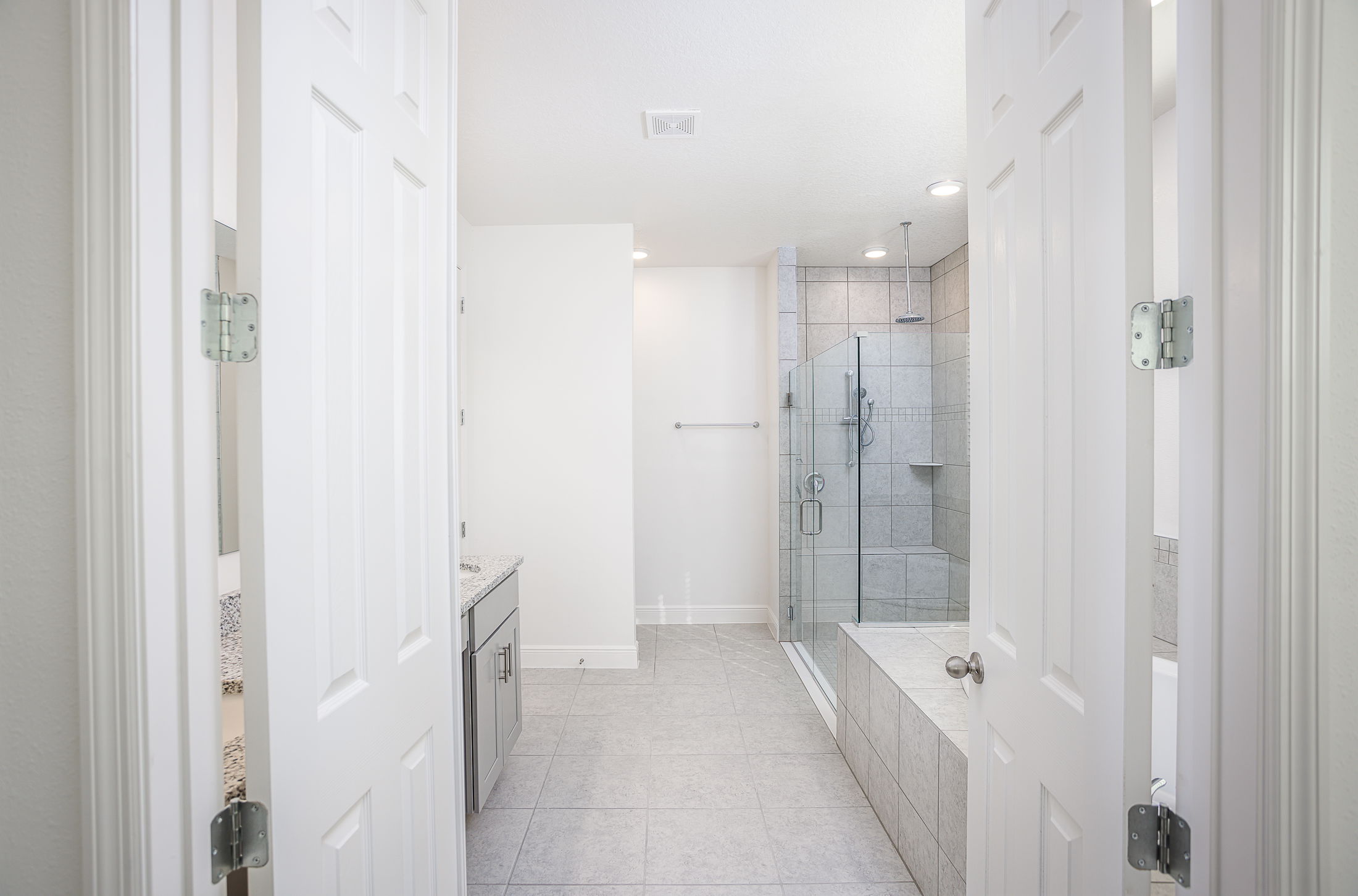 Modern bathroom with glass-enclosed shower, white tile flooring, and granite countertop viewed through open white double doors.