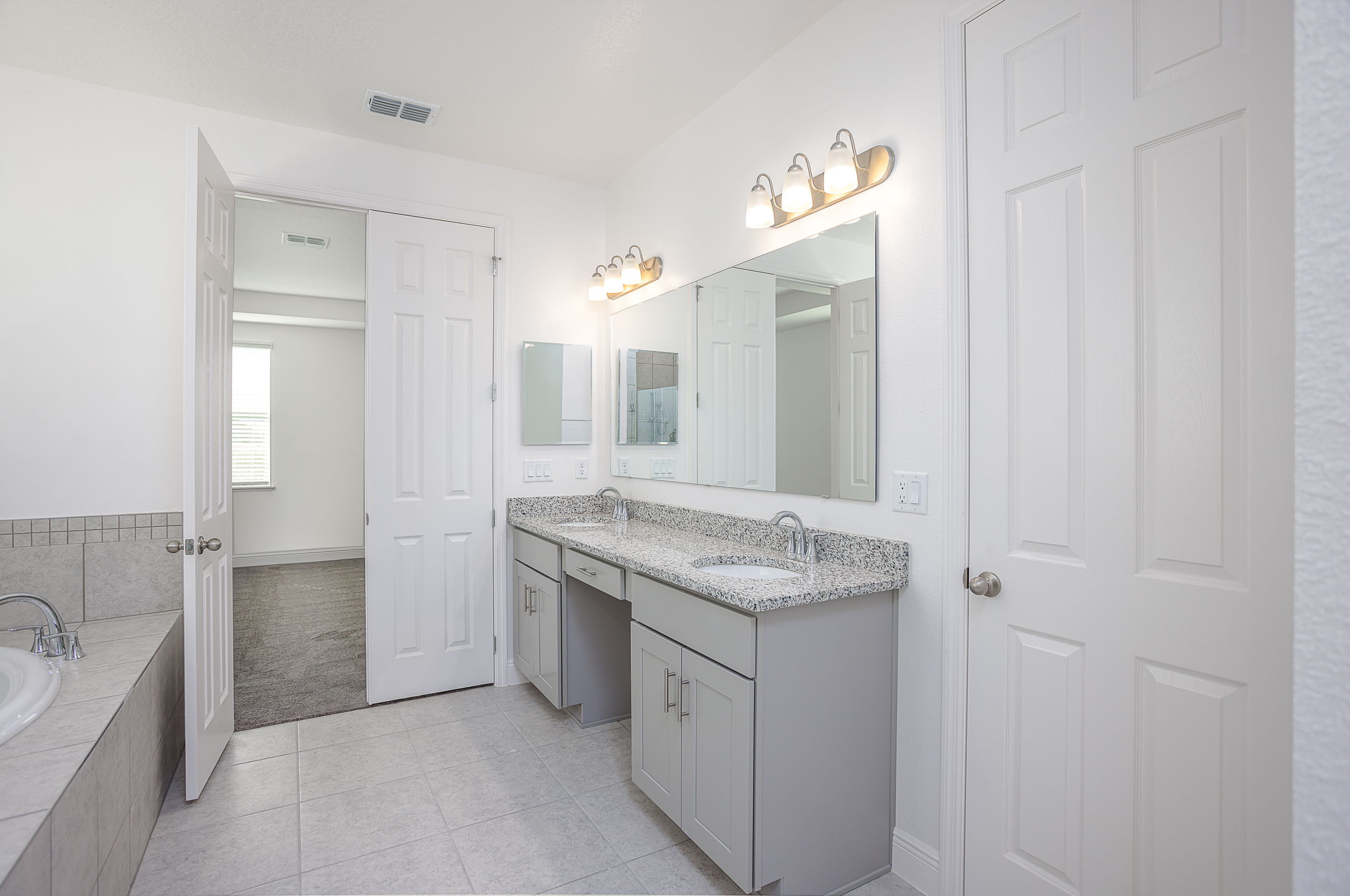 Spacious modern bathroom with dual sink vanity, granite countertops, tiled bathtub, and bright overhead lighting.