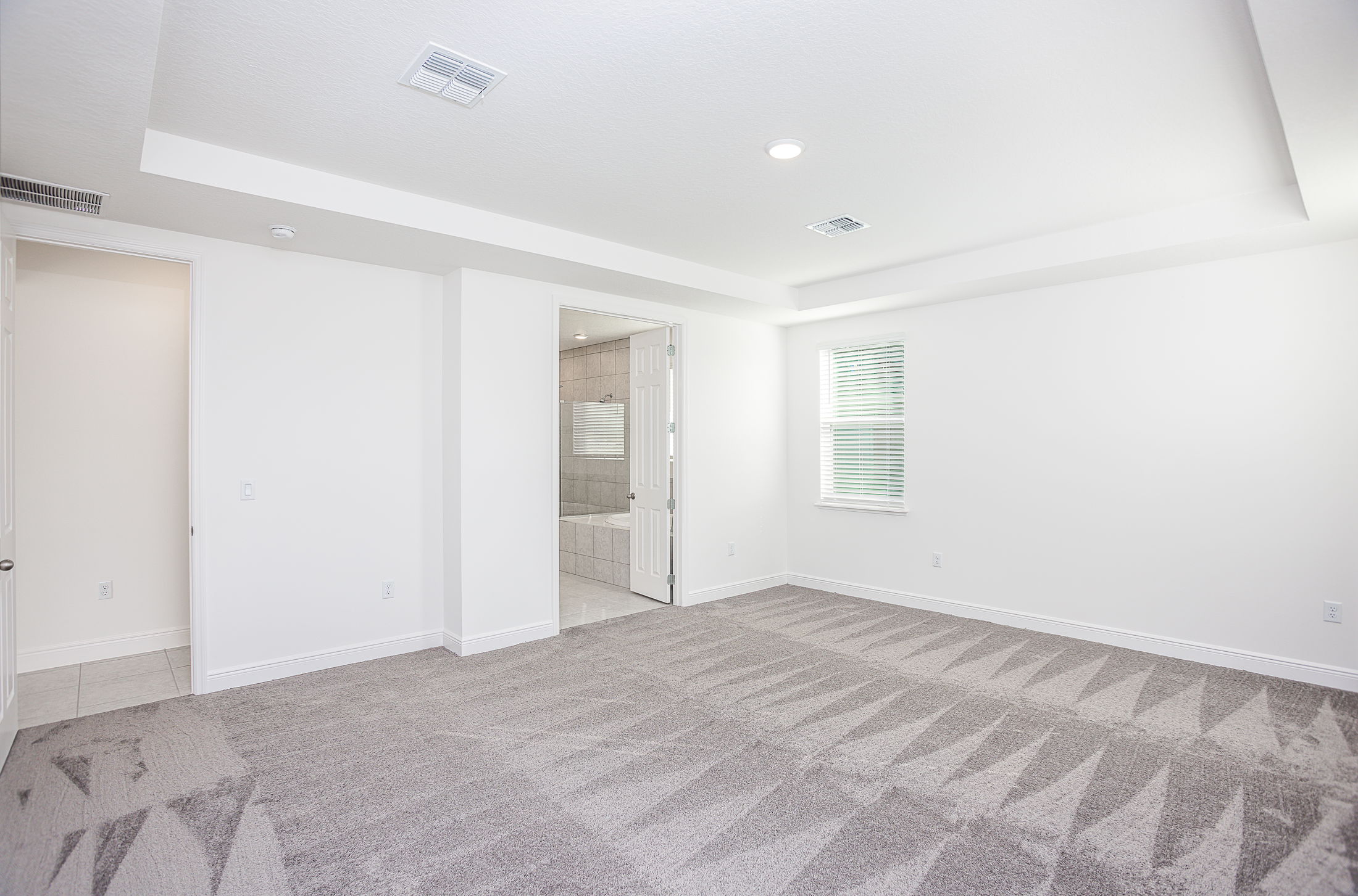 Spacious empty room with freshly cleaned carpet, neutral walls, ceiling lights, and an open doorway leading to a tiled bathroom.