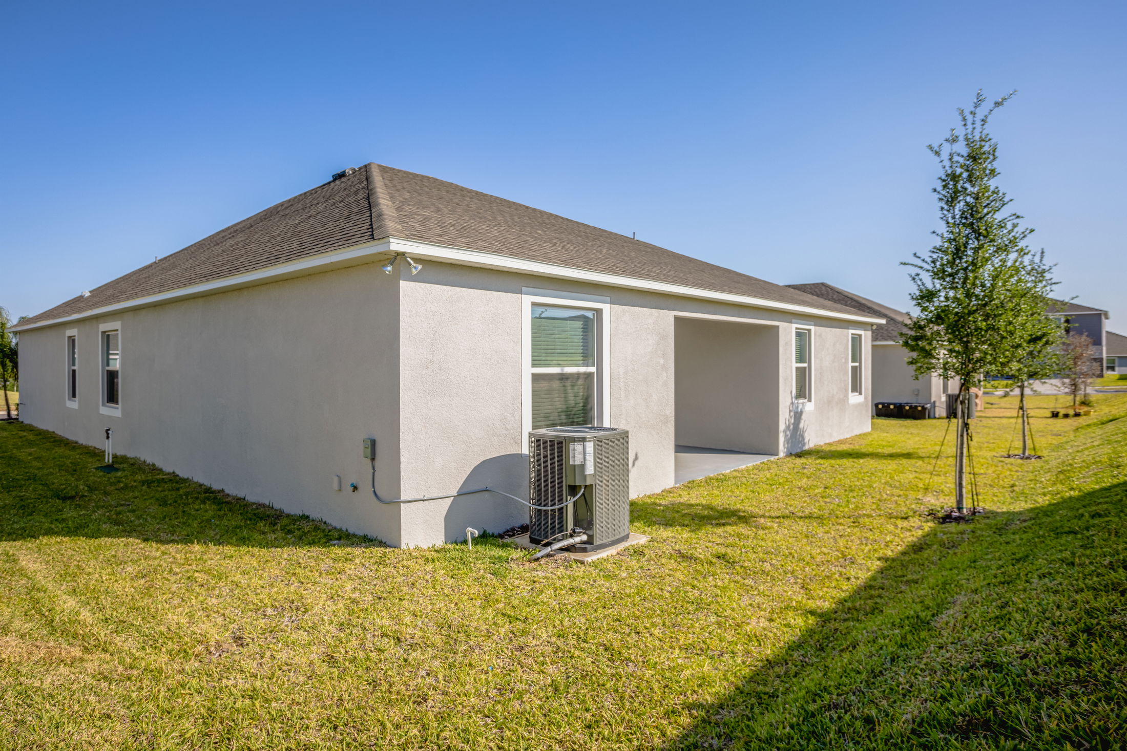 Single-story beige home with a sloped roof, large backyard, and visible air conditioning unit under a clear blue sky.