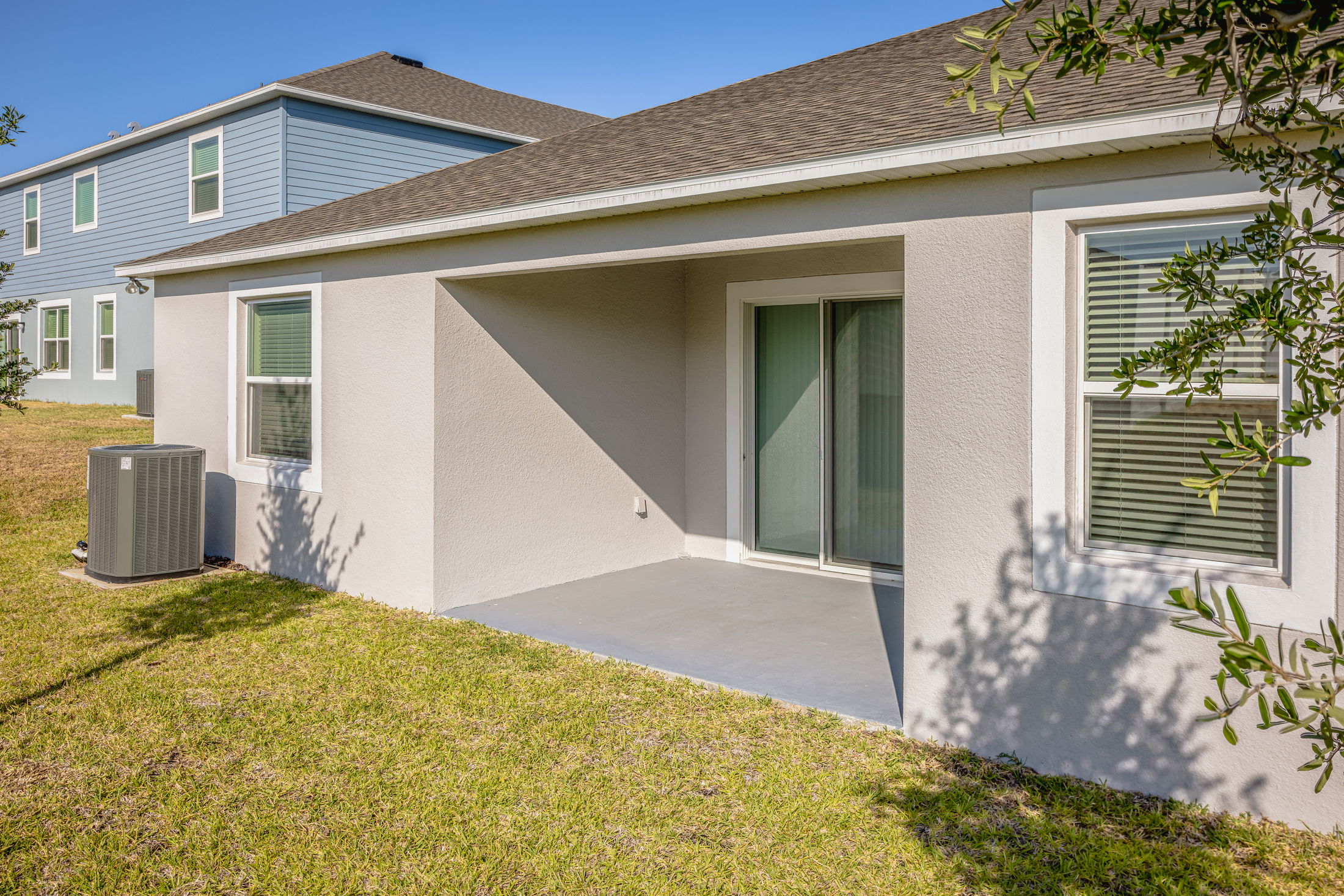 A modern residential backyard with a small covered patio, sliding glass door, adjacent windows, and air conditioning unit on a sunny day.
