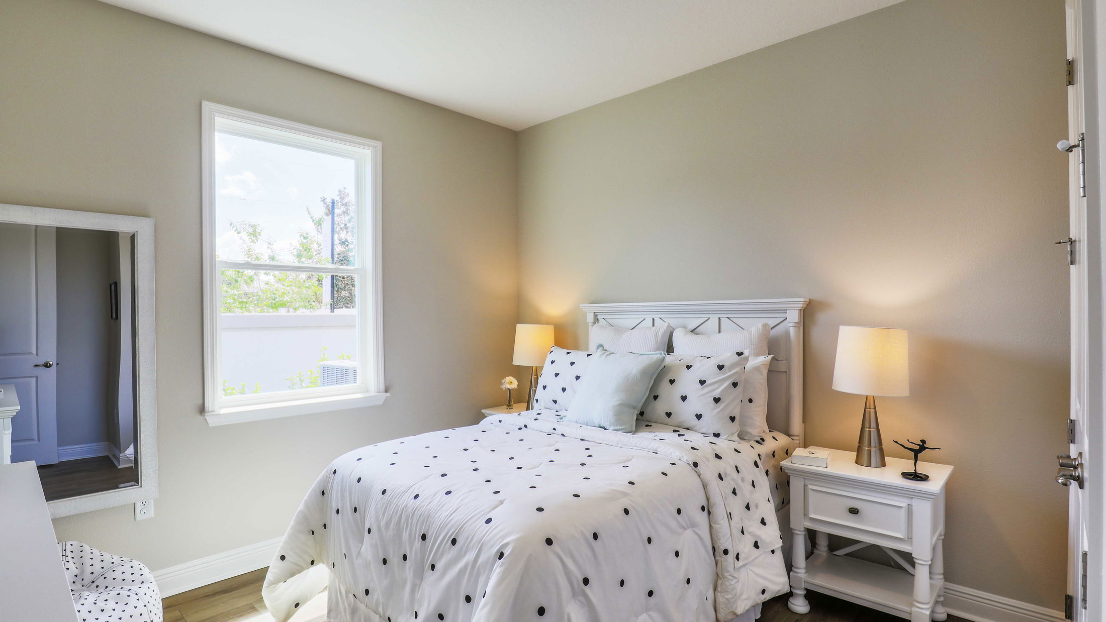 Cozy bedroom with a white polka dot comforter, two bedside tables with lamps, and natural light from a large window.