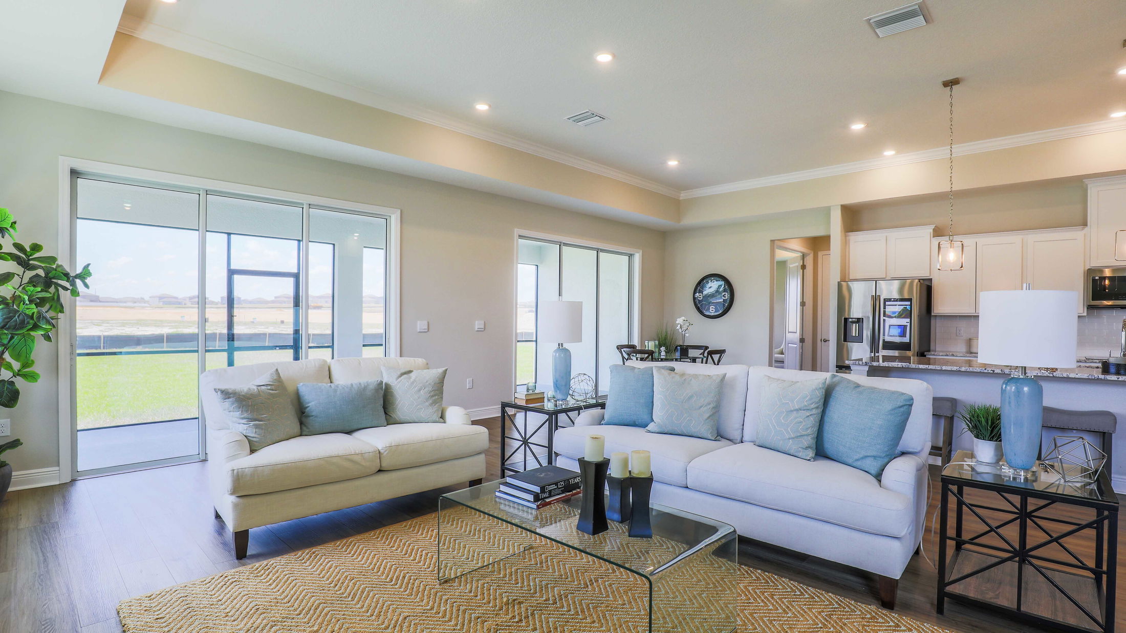 Modern open-concept living room with cozy sofas, glass coffee table, large windows, and view into a sleek kitchen.
