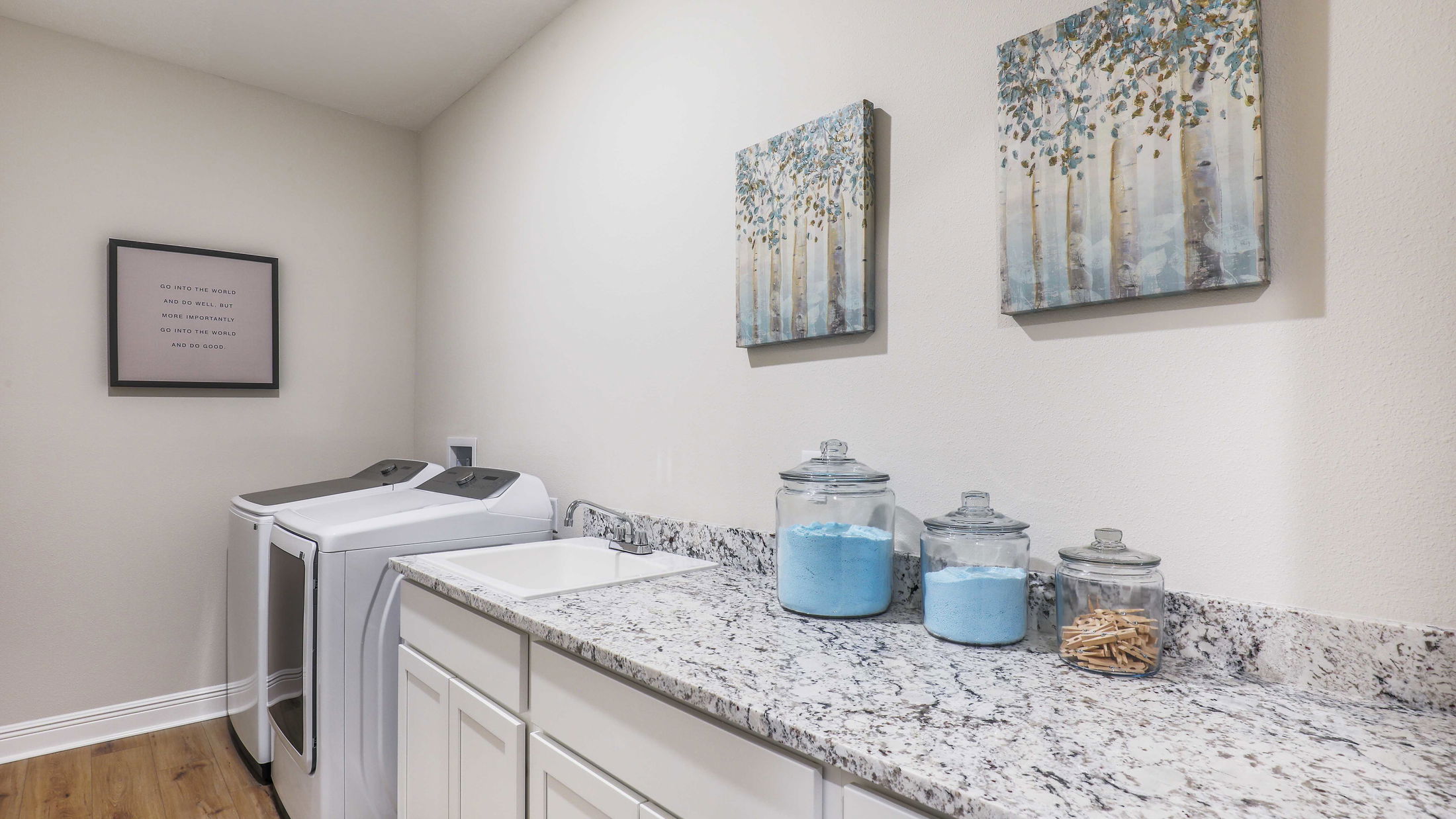 Modern laundry room with a granite countertop, washing machine, jars of laundry supplies, and framed wall art.