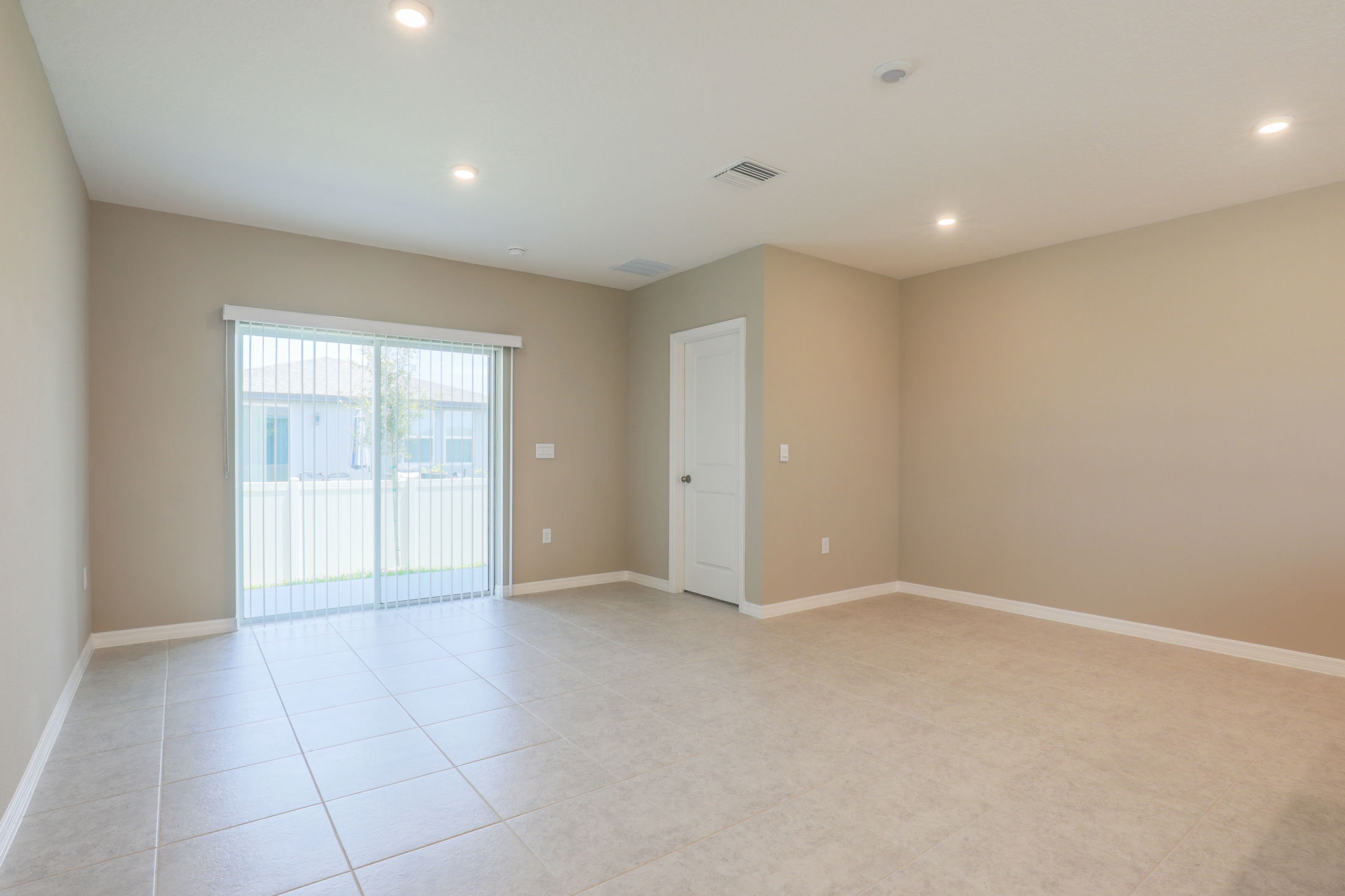 Empty beige room with tiled floor, recessed lighting, sliding glass doors, and a closed white door.