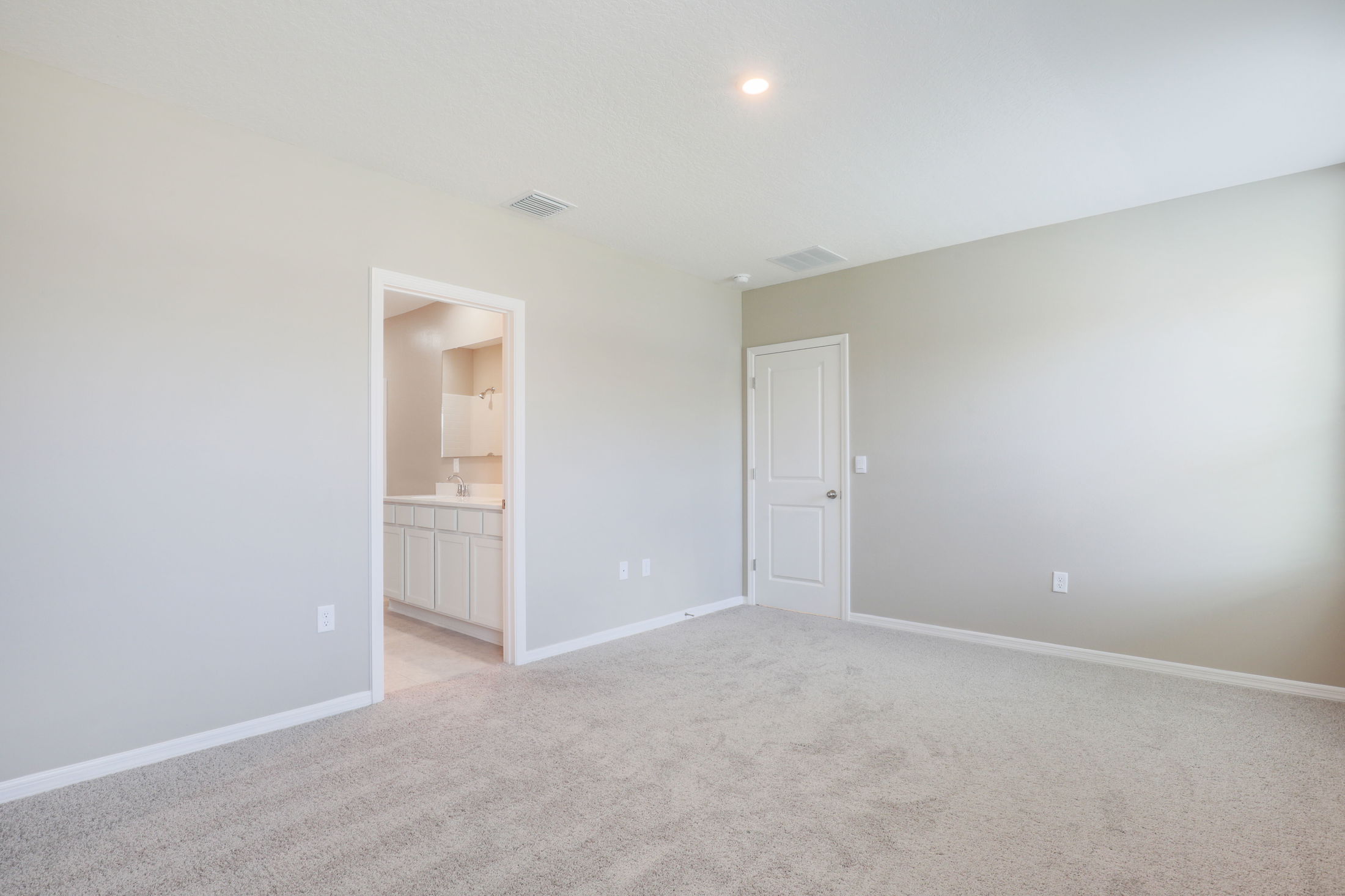 Spacious empty room with beige carpet, closed white door, and visible bathroom entrance.