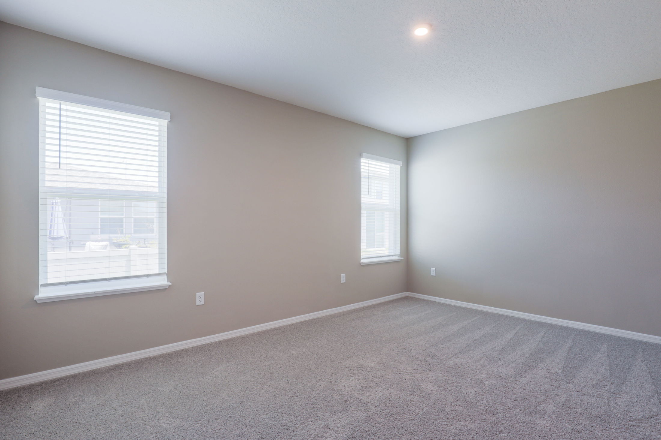 A bright, empty room with beige walls, carpeted floor, and two windows with blinds letting in natural light.