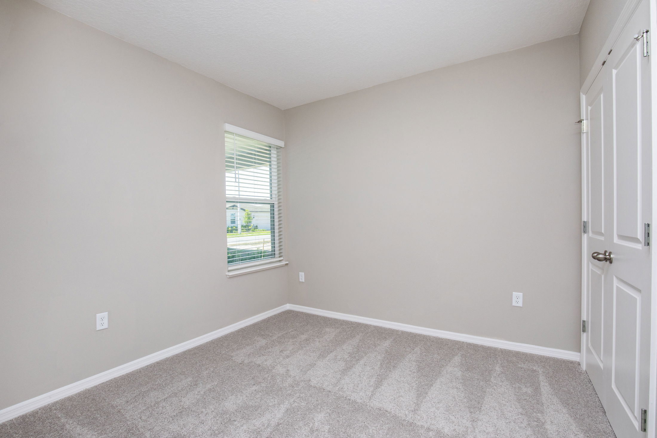 Empty room with beige walls, carpeted floor, a window with blinds, and a closed door, providing a bright and clean interior space.
