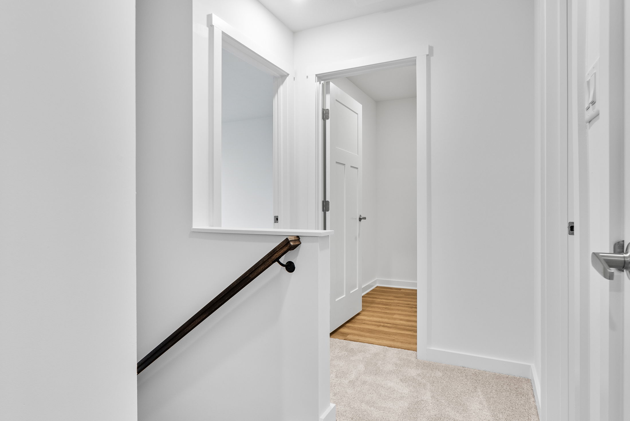 A modern interior hallway with white walls, wooden flooring, a carpeted area, and a dark wooden handrail.