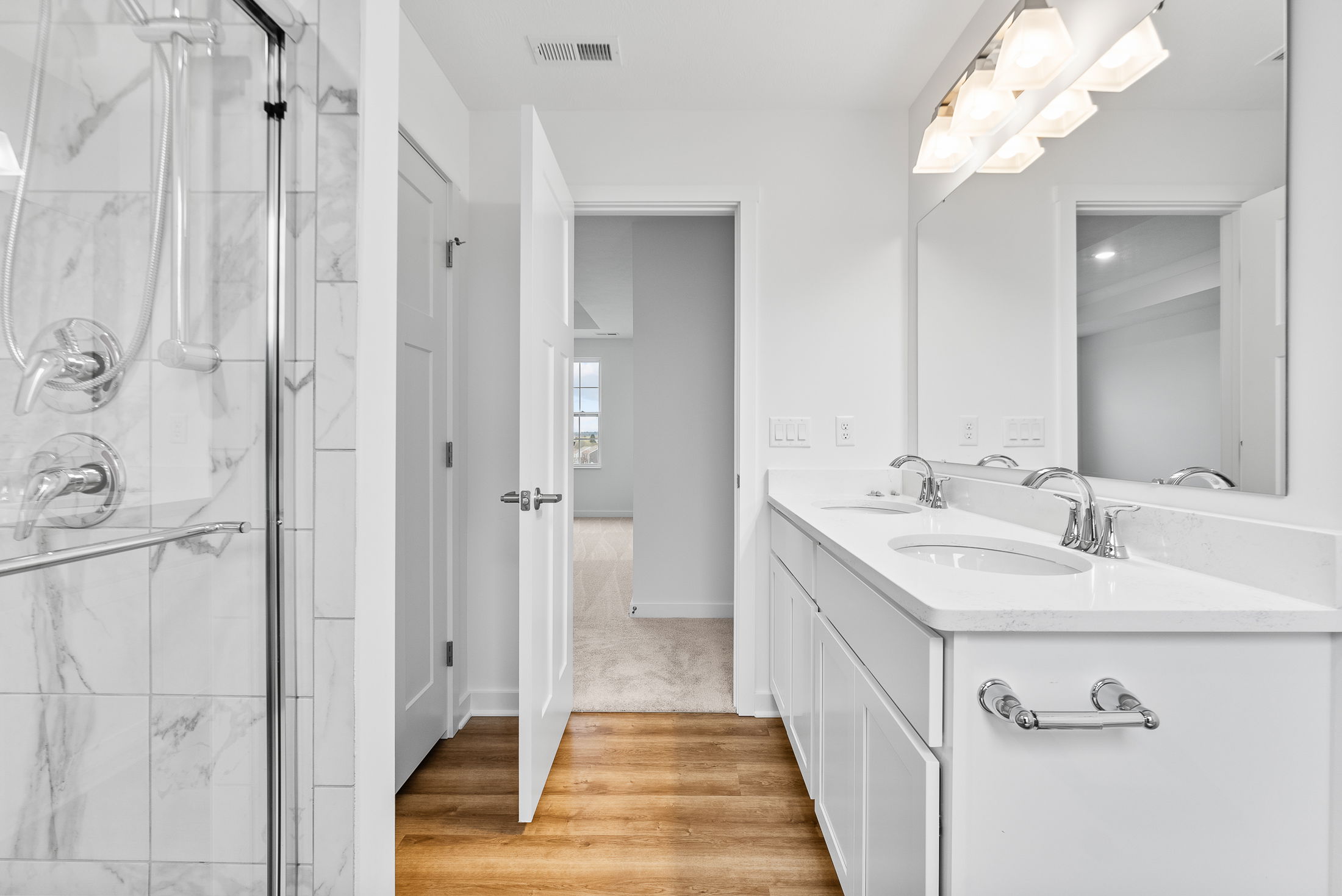 Modern white bathroom with dual sinks, marble shower, and hardwood floors leading to an open door.