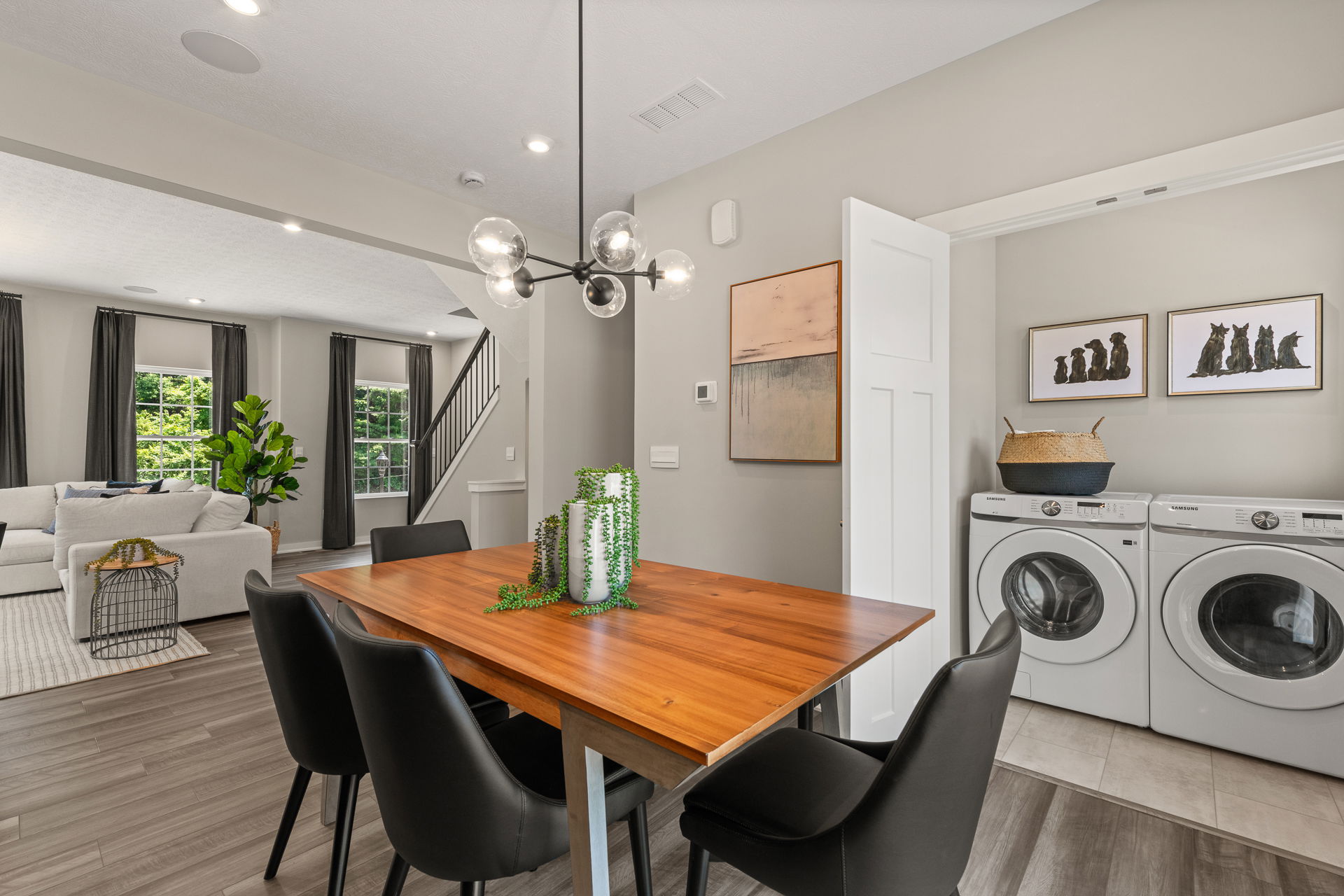 Modern dining area with a wooden table, black chairs, contemporary chandelier, and an adjacent laundry nook featuring Samsung washer and dryer.