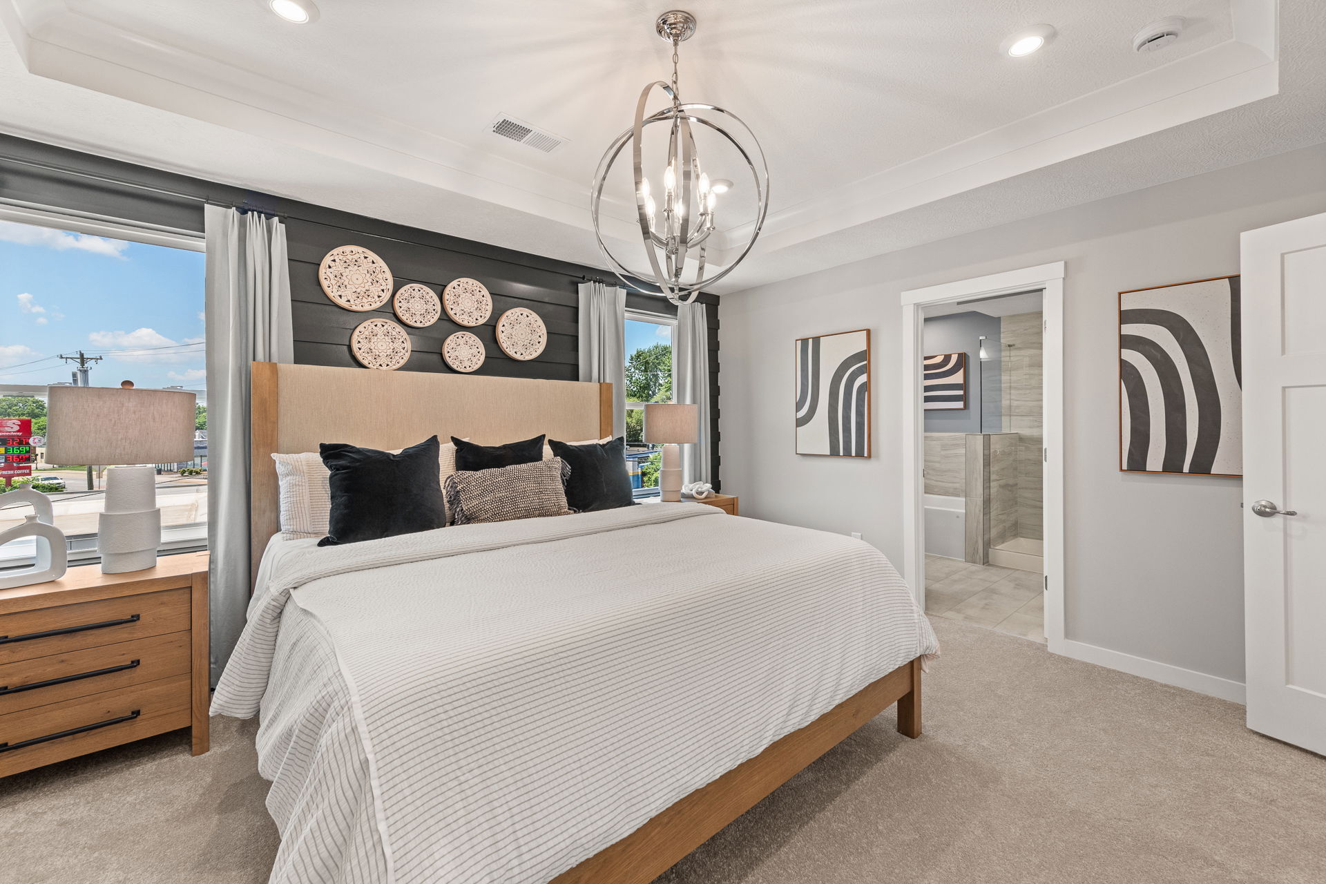 Modern bedroom featuring a beige bed, decorative wall plates, contemporary art, and a stylish chandelier near a large window.