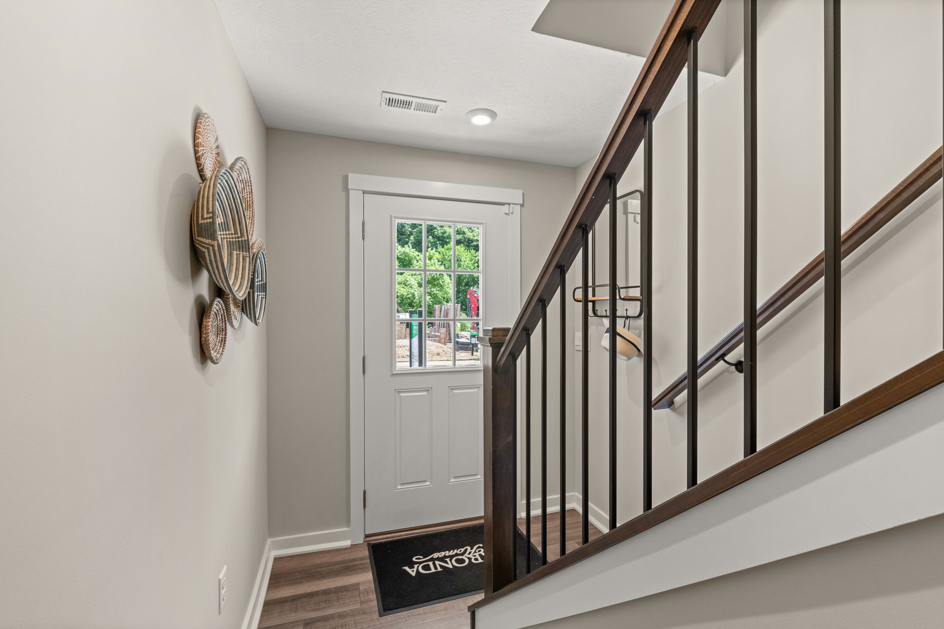 Modern entryway with staircase, wicker wall decor, and a white door leading to a lush green garden.