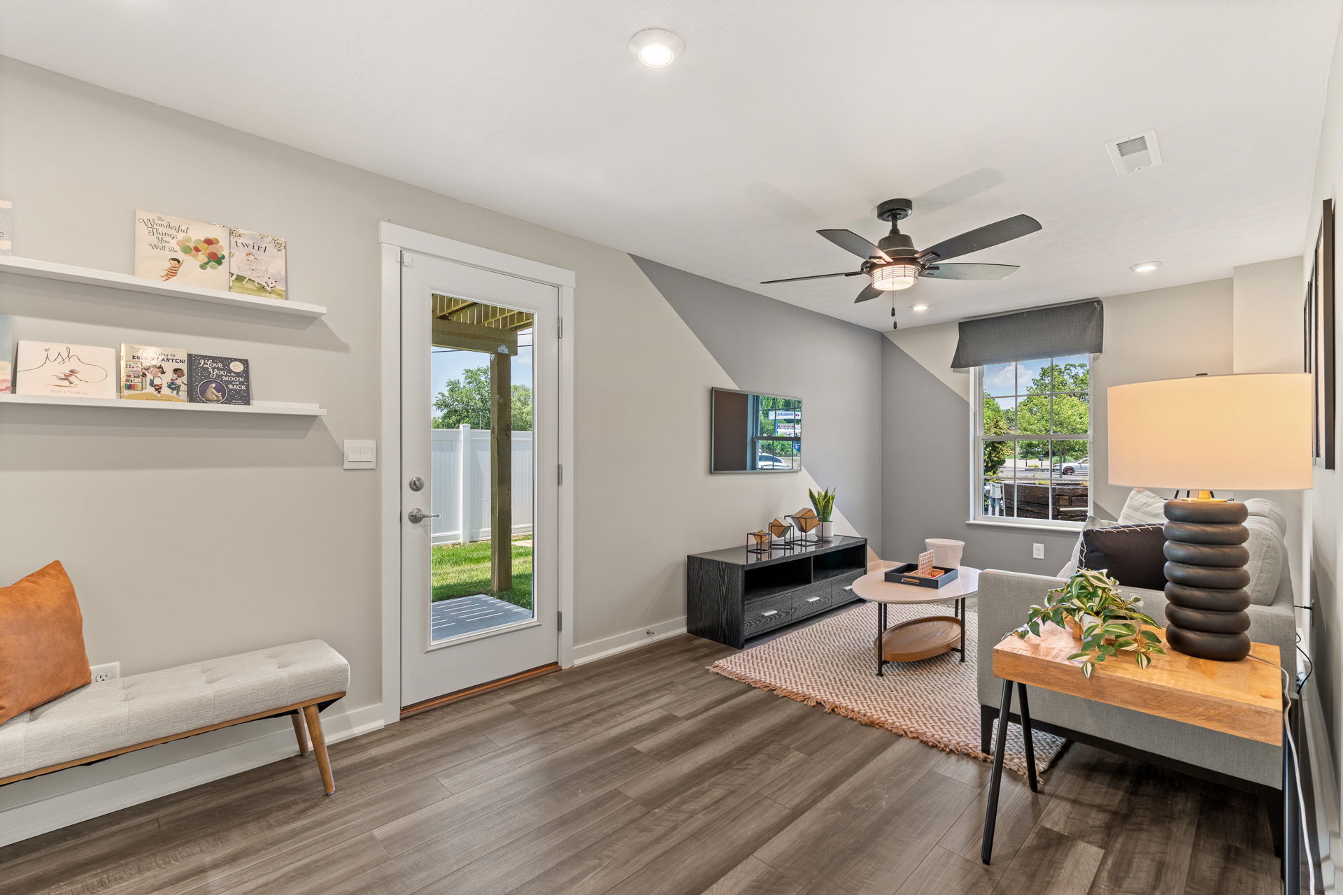 Modern living room with a ceiling fan, gray sofa, wall-mounted TV, and large window overlooking a backyard.