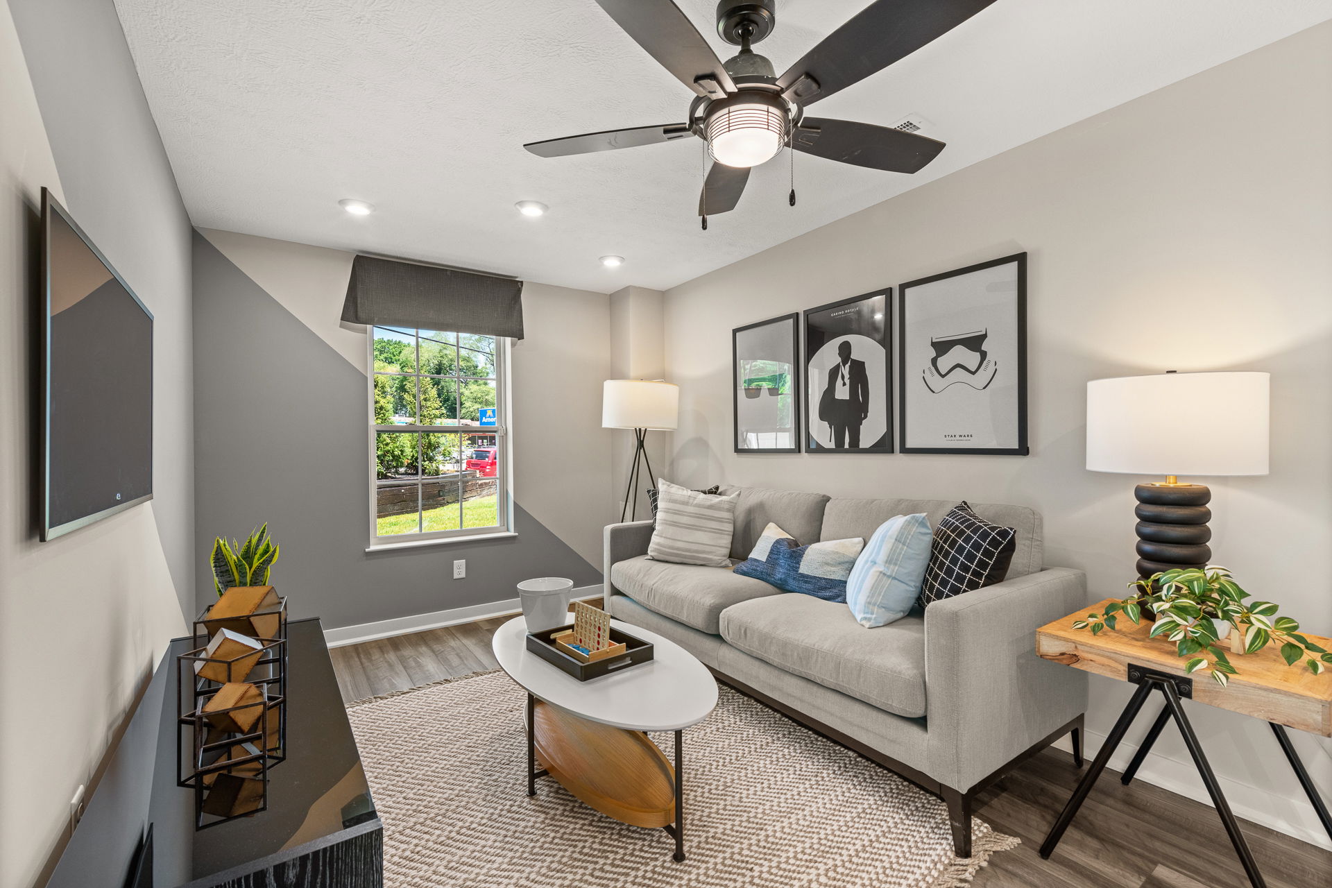 Modern living room with a gray sofa, stylish artwork, contemporary lighting, ceiling fan, and a view of the outdoors through a large window.