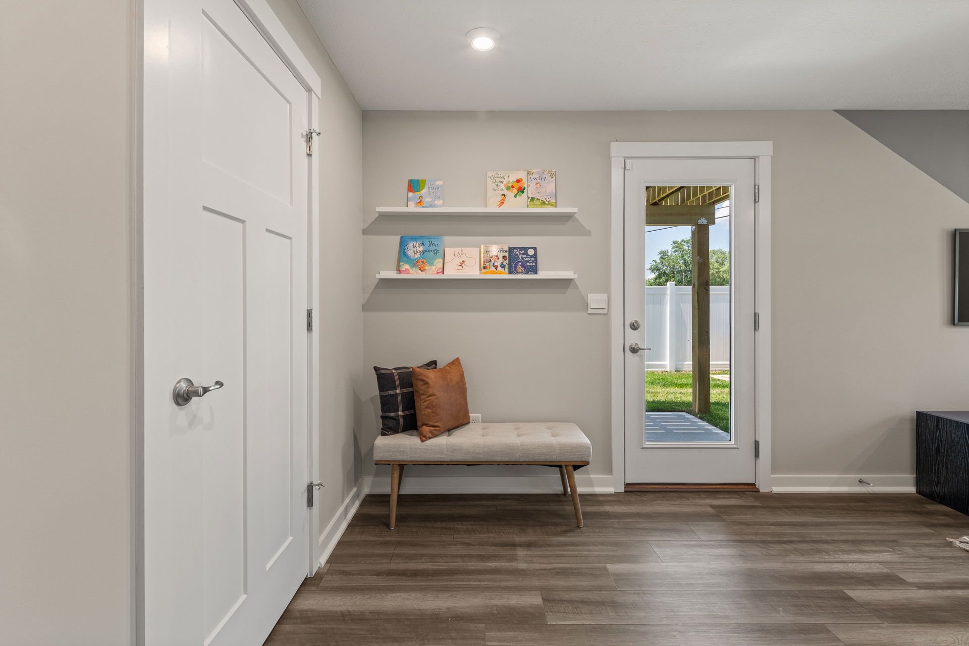 Modern entryway interior with a white door, minimalist bench, decorative pillows, floating shelves with books, and glass door to outdoor space.
