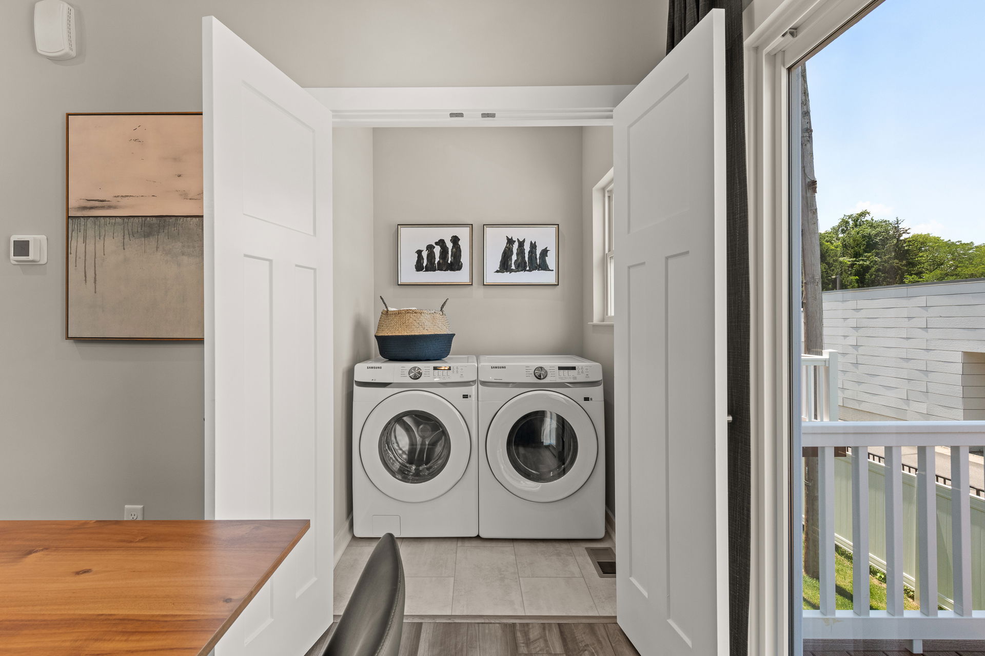 A modern laundry room with a Samsung washer and dryer, decorative basket, and framed artwork, adjacent to a wooden desk and window view.